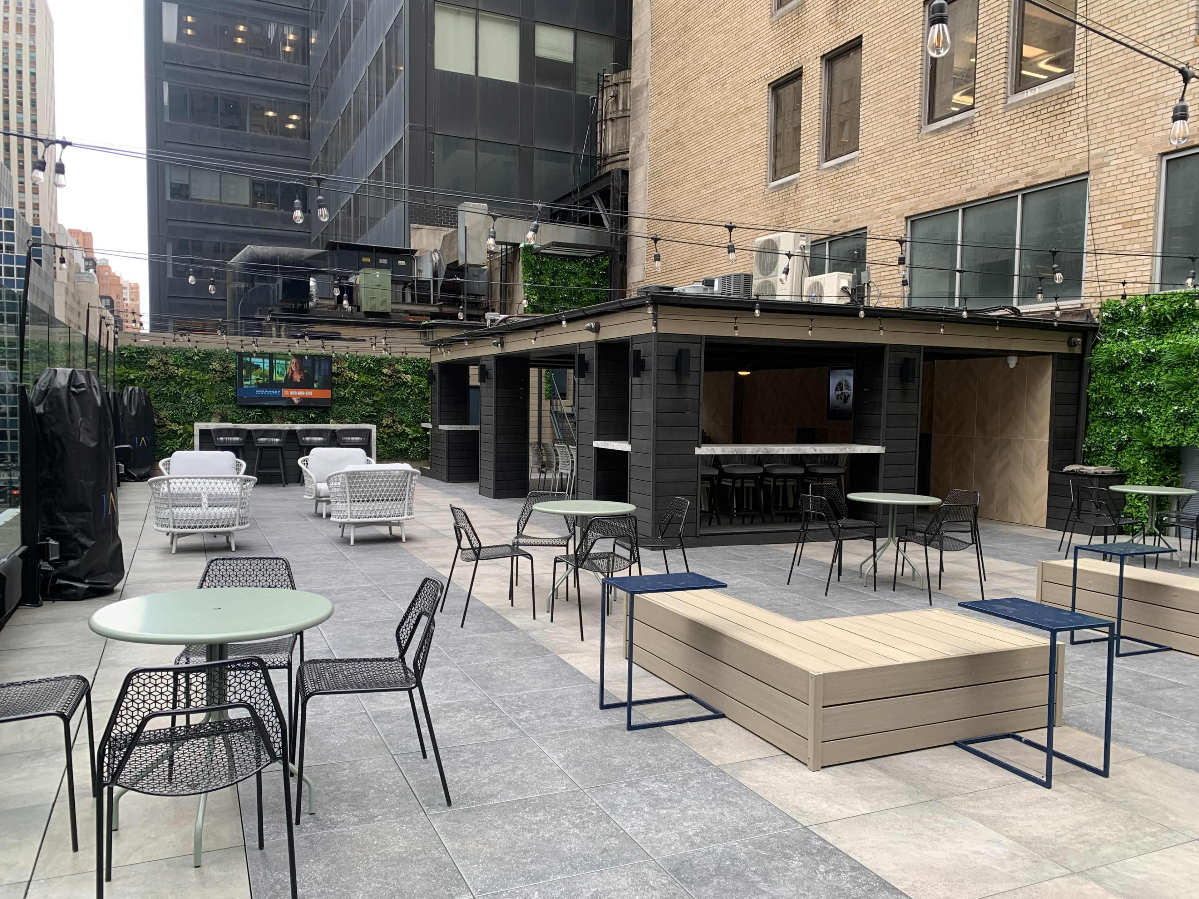 The image shows a rooftop terrace equipped with several tables and seating areas, surrounded by greenery and buildings.