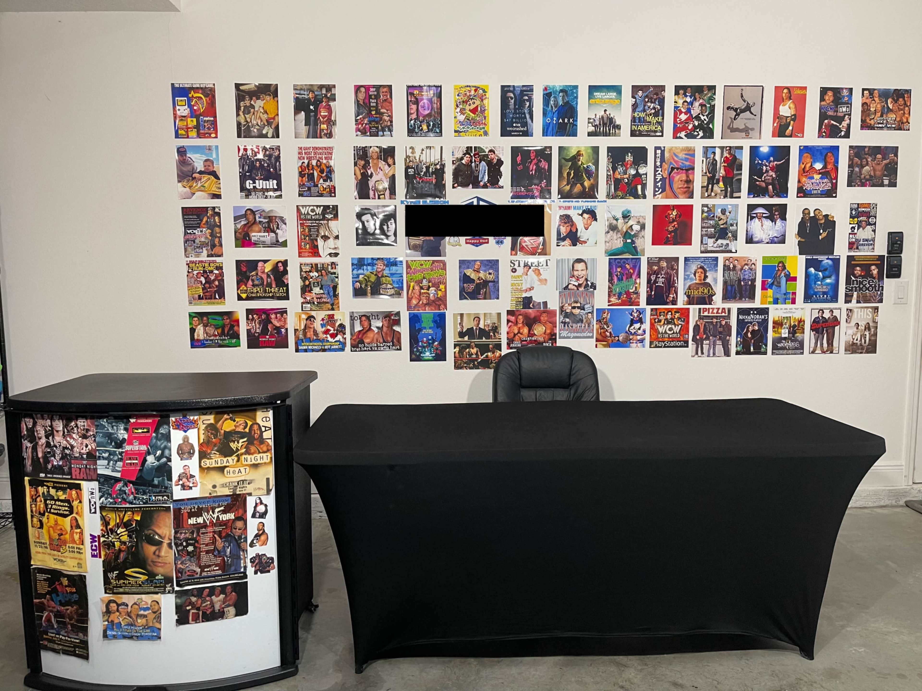 The image shows a setup featuring two tables, one covered with a black tablecloth and the other displaying posters on its front, against a wall adorned with a variety of colorful posters.