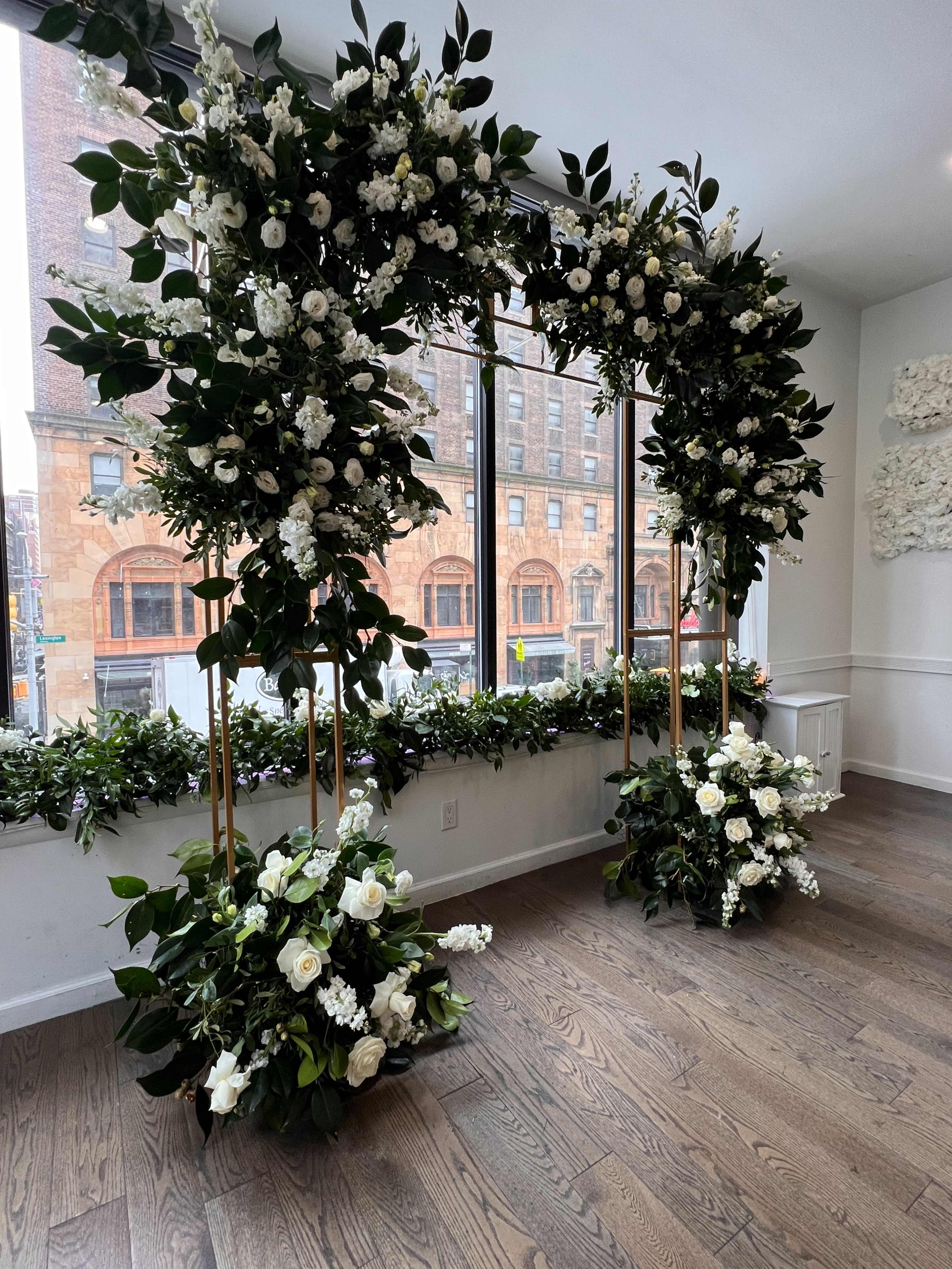 The image shows two floral arrangements with white flowers and greenery arranged in a doorway, positioned against a backdrop of large windows overlooking a city street.