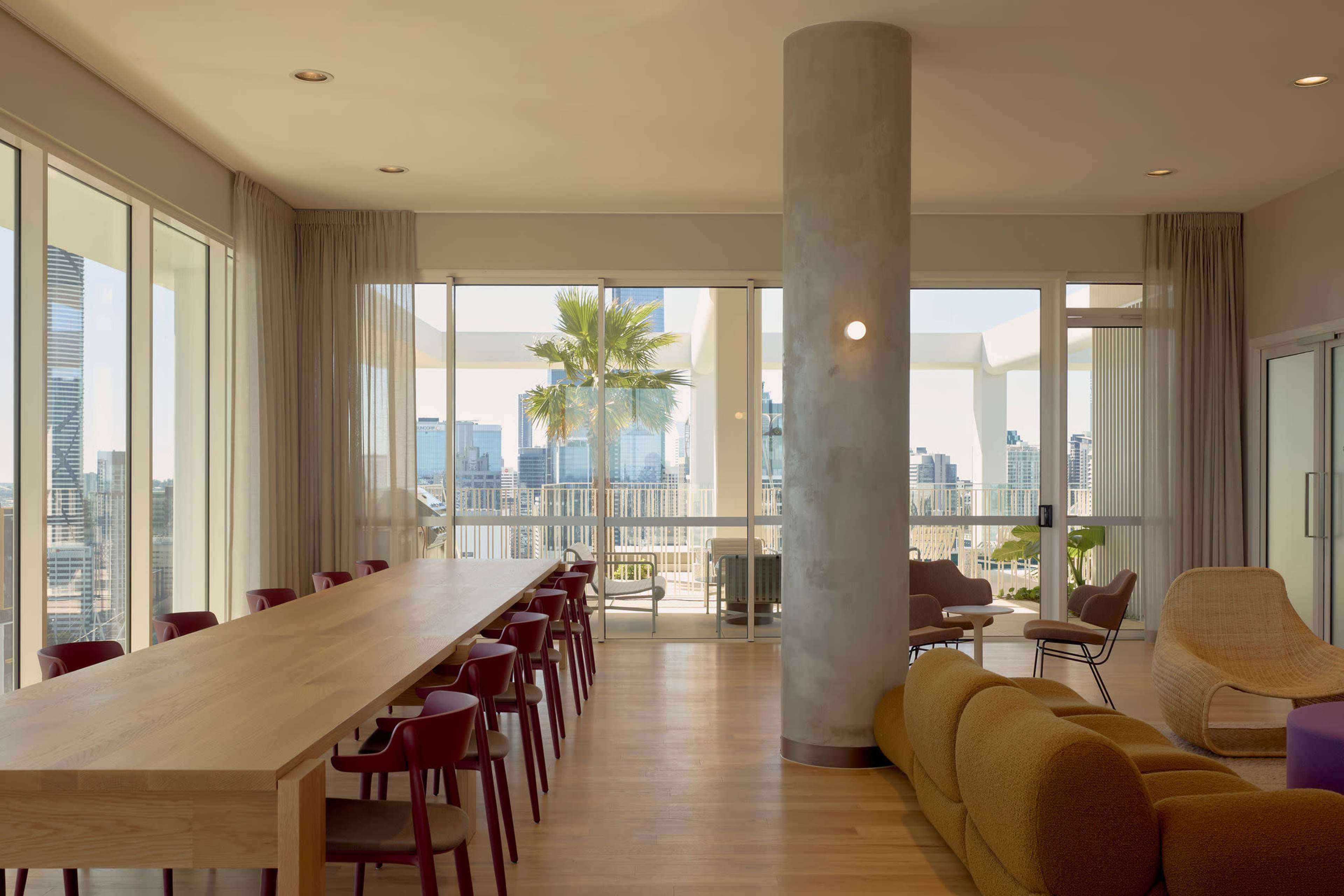A modern, open-concept room features a long wooden dining table, colorful chairs, and large windows offering a view of a city skyline with palm trees outside.