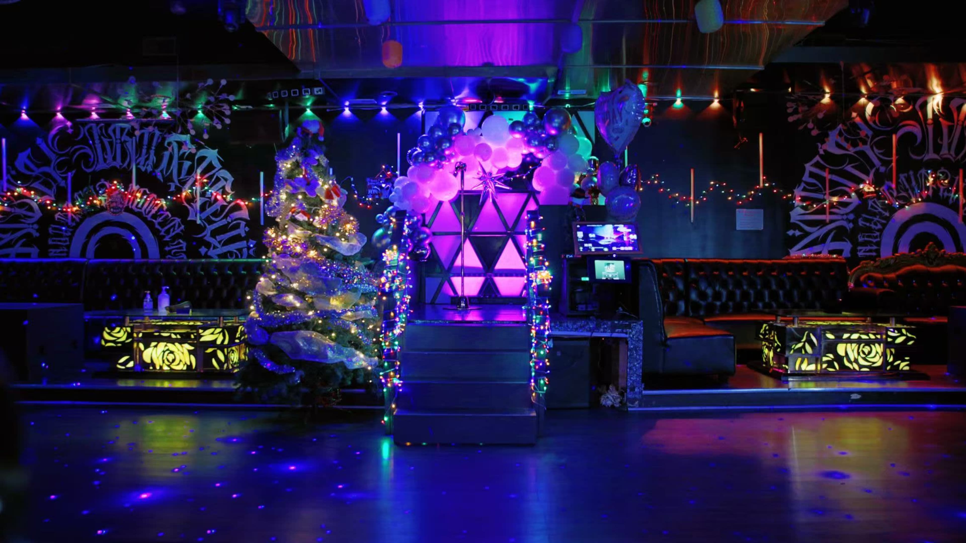 The image shows a dimly lit nightclub with a decorated Christmas tree in one corner and a stage featuring colorful lights and a geometric structure at the center.