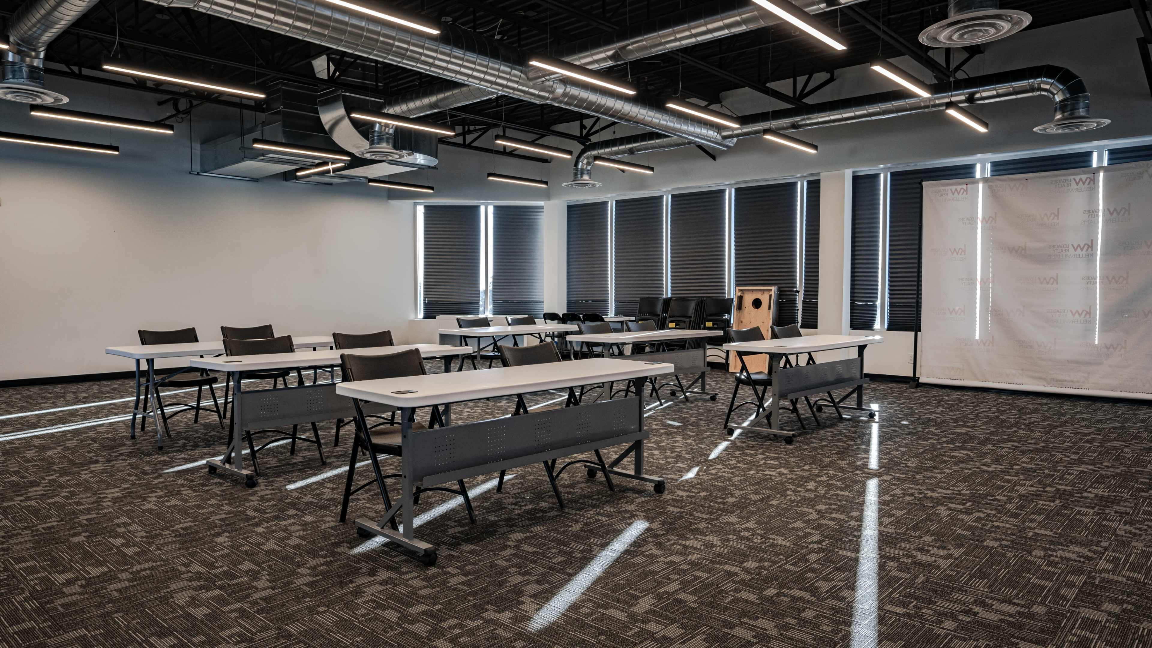 A modern classroom features several folding tables and chairs arranged under bright overhead lights, with large windows partially covered by shades.