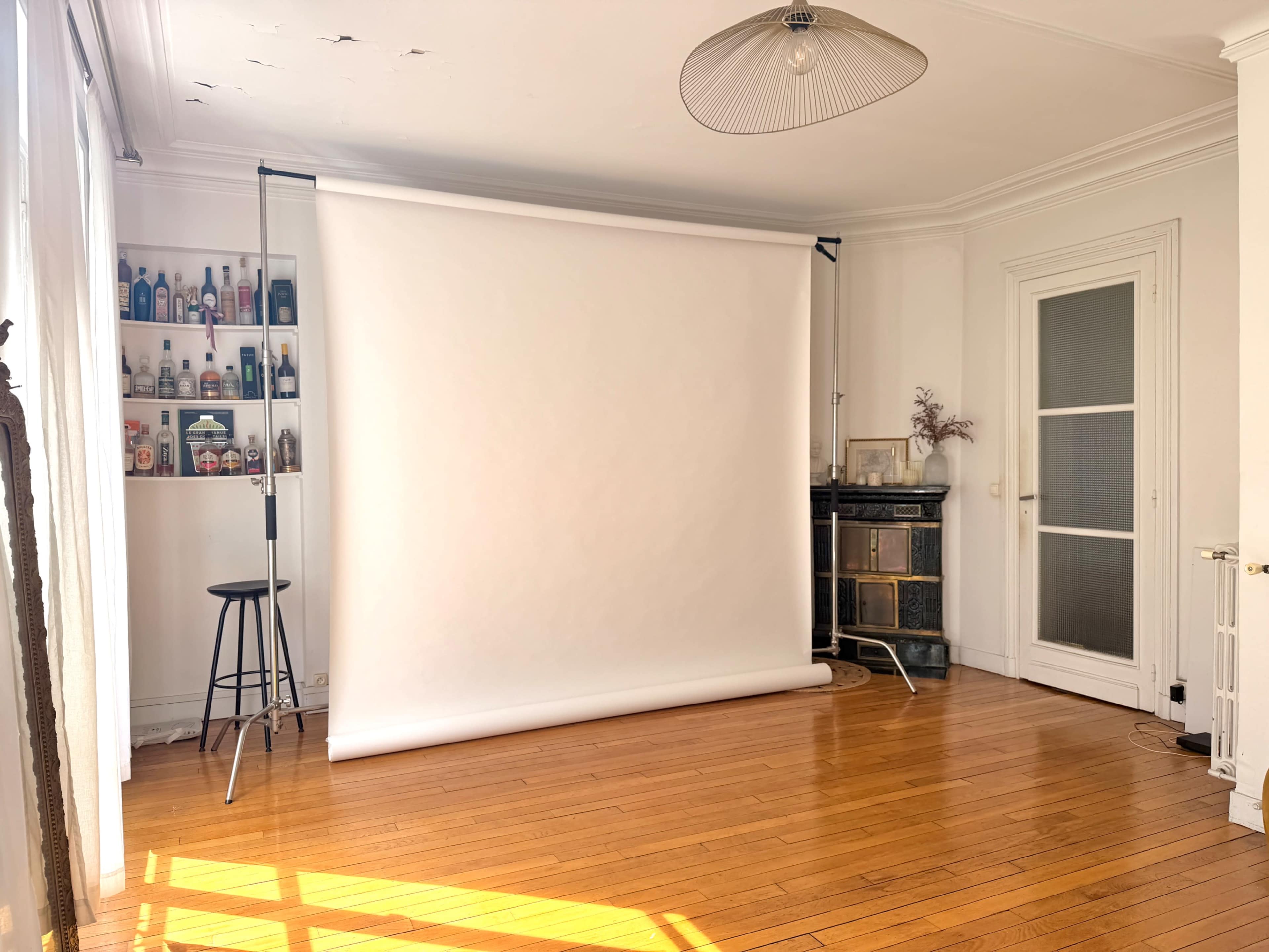 The image shows a spacious, well-lit room featuring a blank white backdrop on a stand, a bar stool, and a shelf with various beverage bottles.
