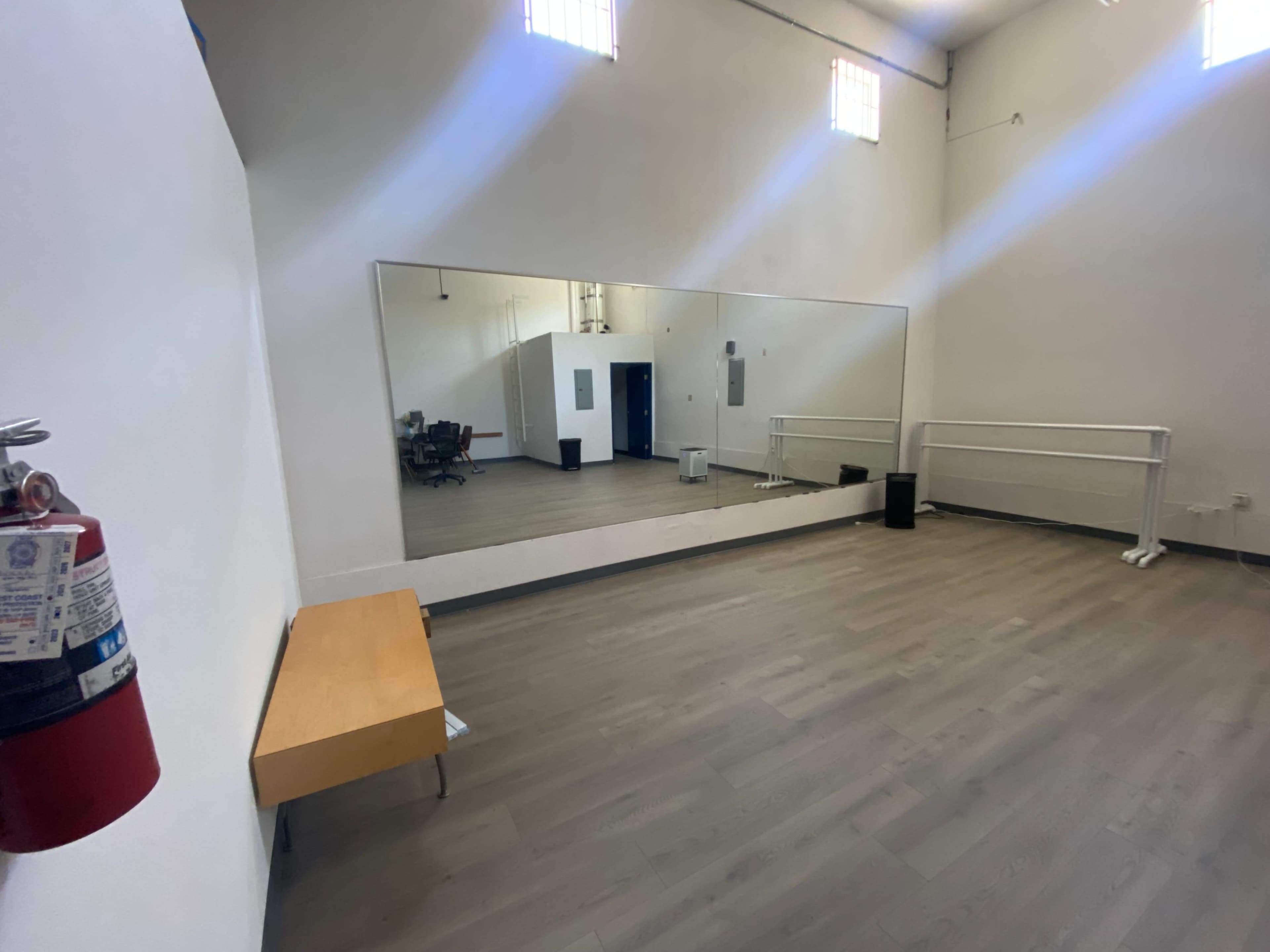 A spacious dance studio with a large mirror, ballet barre, wooden bench, and fire extinguisher on the wall.