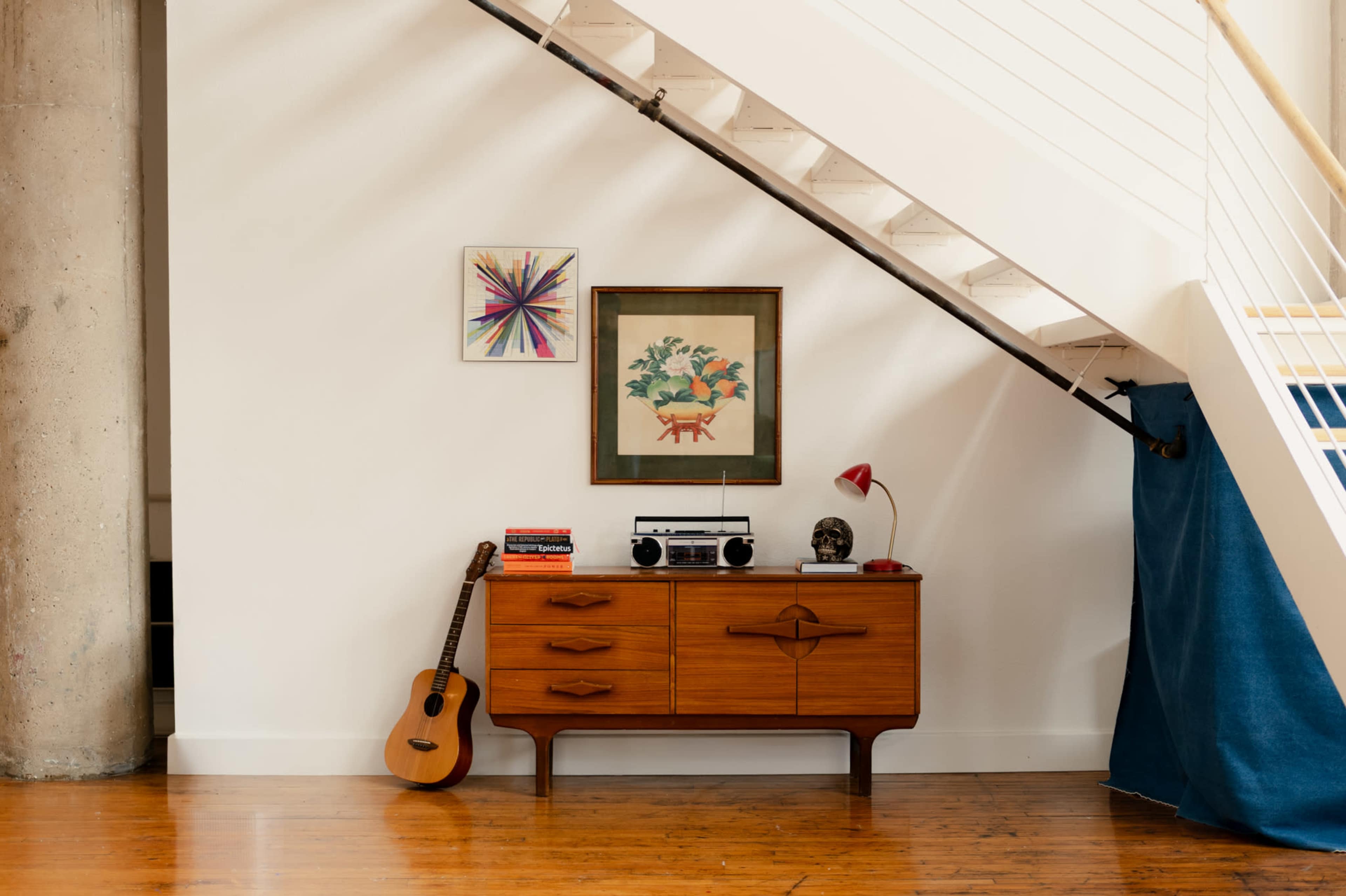 A wooden sideboard with a vintage radio and a lamp is placed under a staircase, accompanied by a guitar and art on the wall.