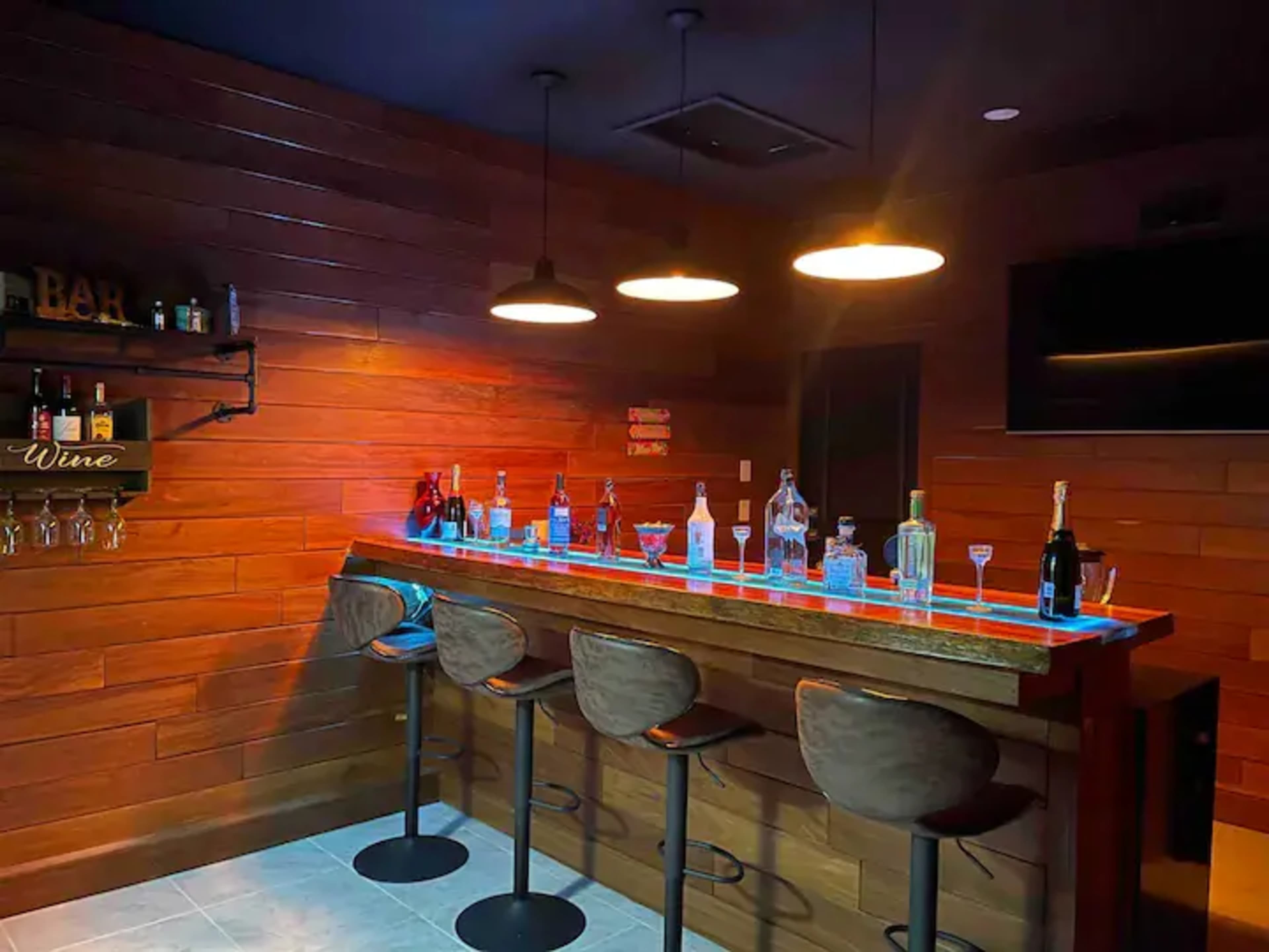A modern home bar with a wooden interior, illuminated by pendant lights, featuring a long bar counter and several bottles of liquor displayed on the shelf and countertop.