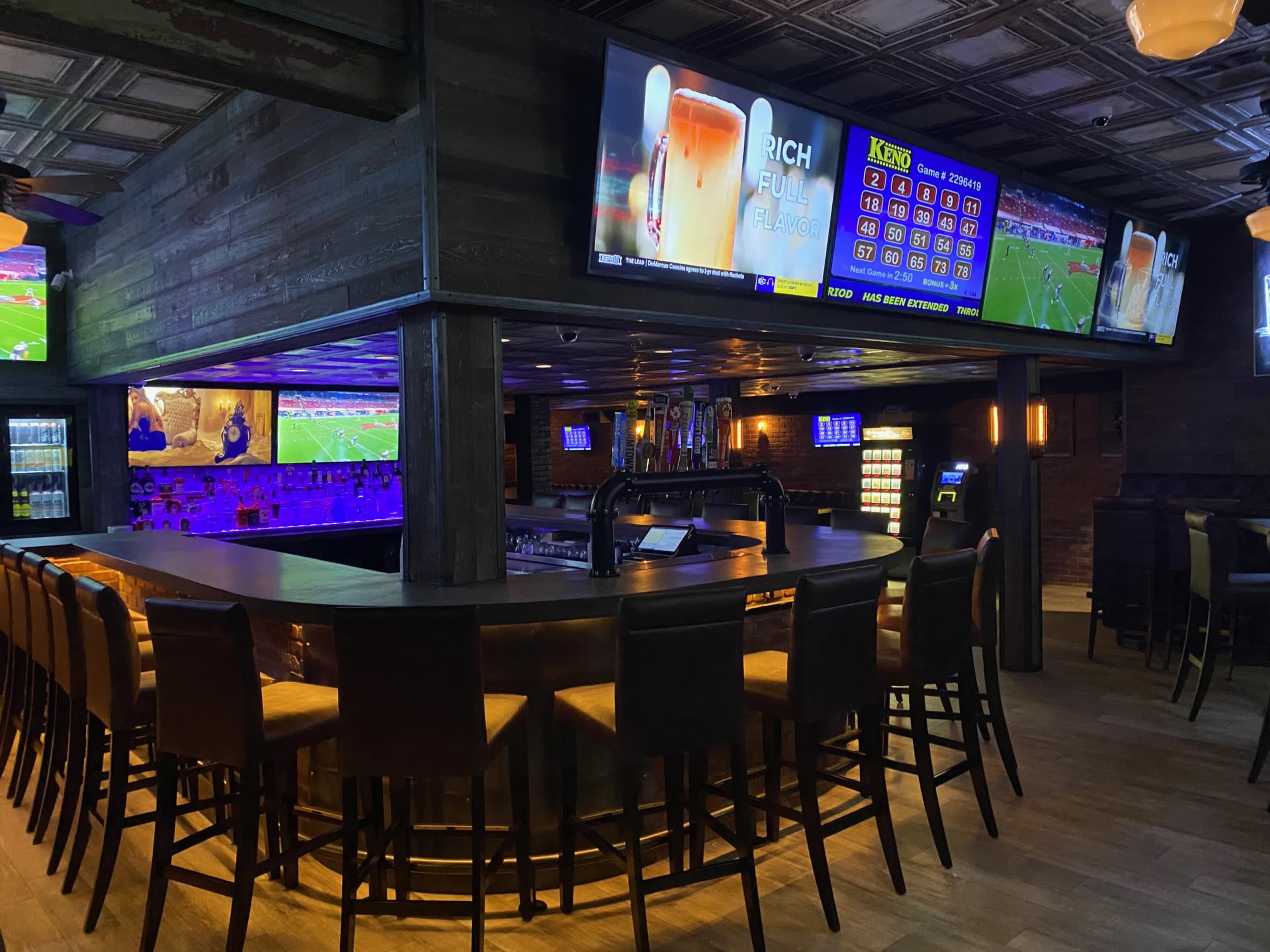 The image shows a dimly lit bar with high stools, multiple large screens displaying sports events, and a curved counter lined with bottles.