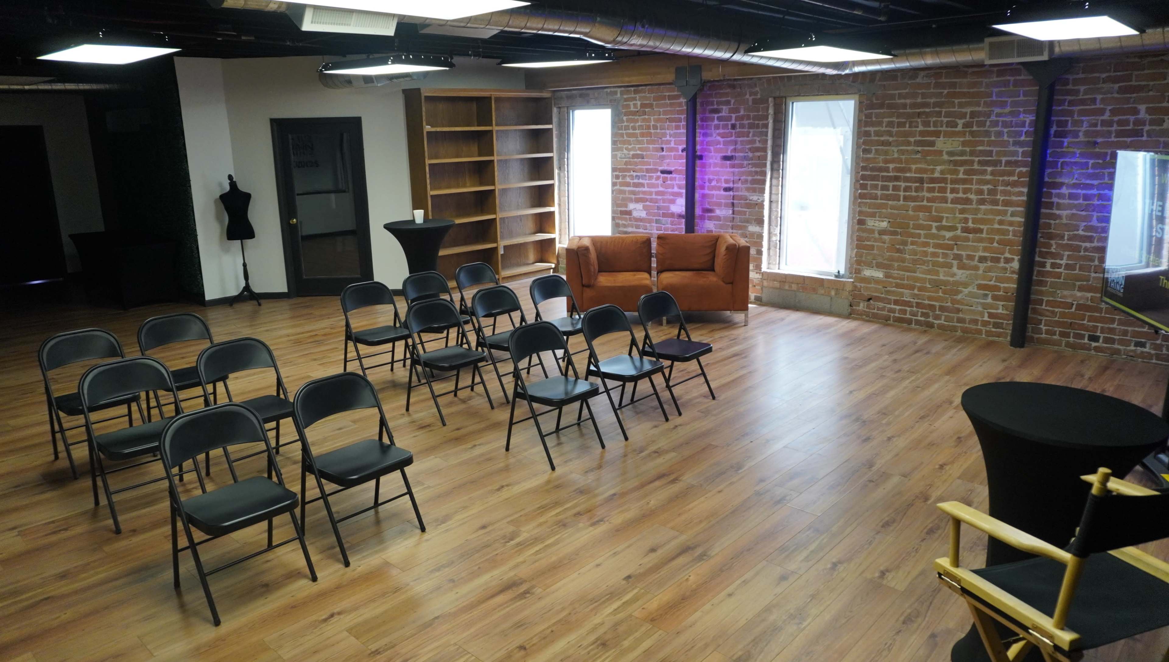 A spacious room with wooden flooring, featuring several black chairs arranged in rows, a brown couch against a brick wall, and a few large windows allowing natural light.