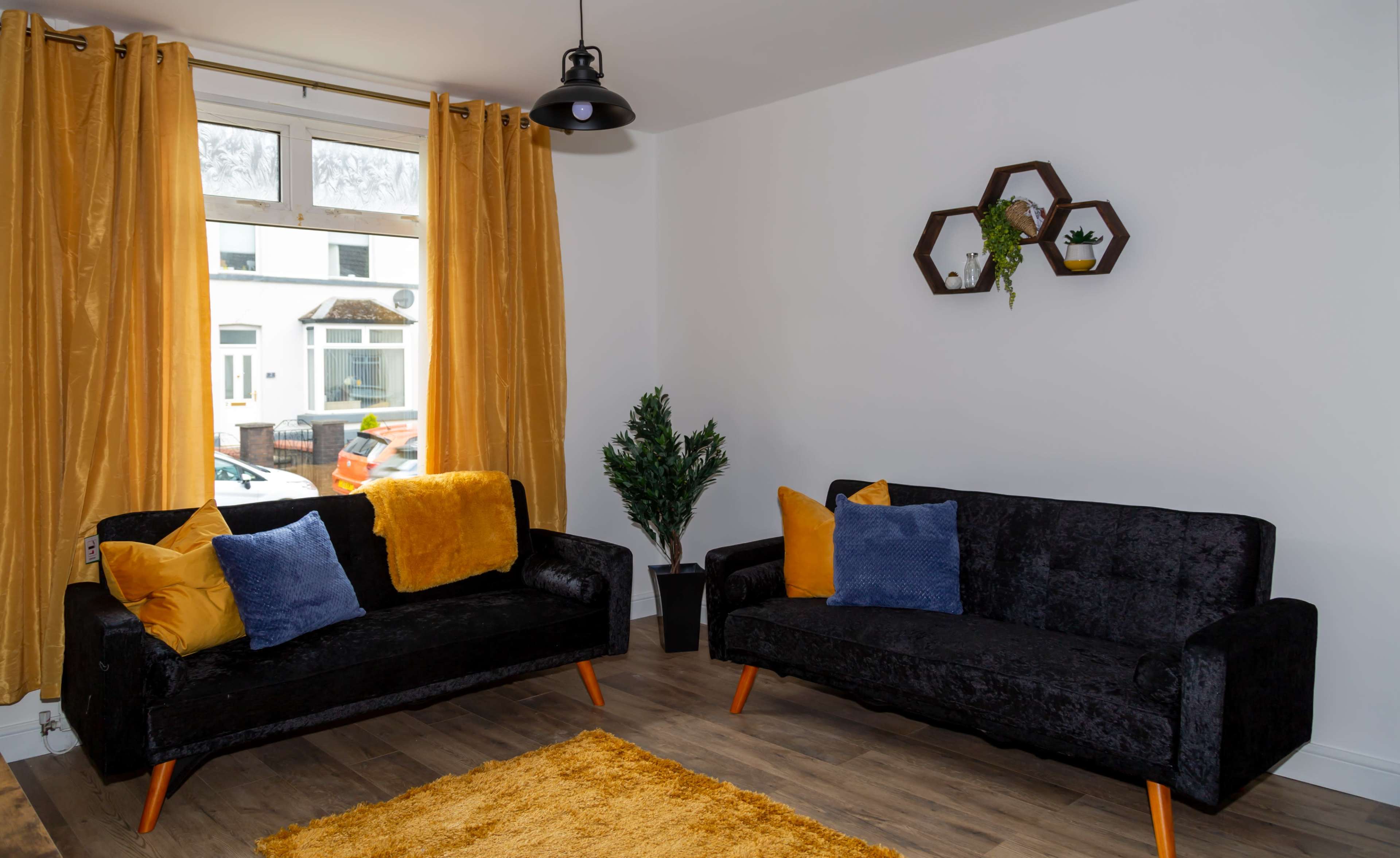 A living room features two black sofas with colorful cushions, a yellow rug on the wooden floor, and plants on the wall.