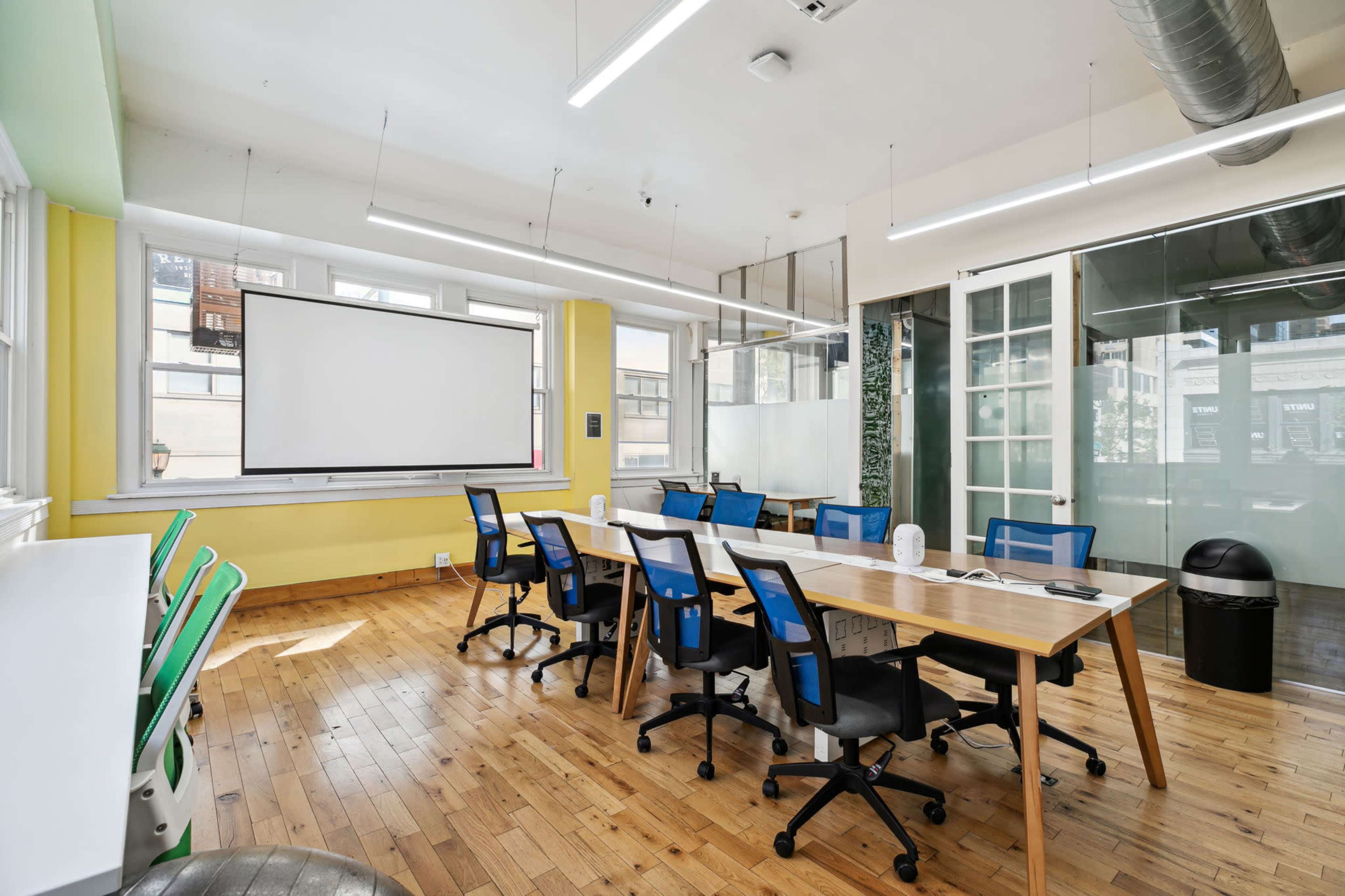 A modern office space features several workstations with blue chairs, a large screen mounted on the wall, and a green accent wall.