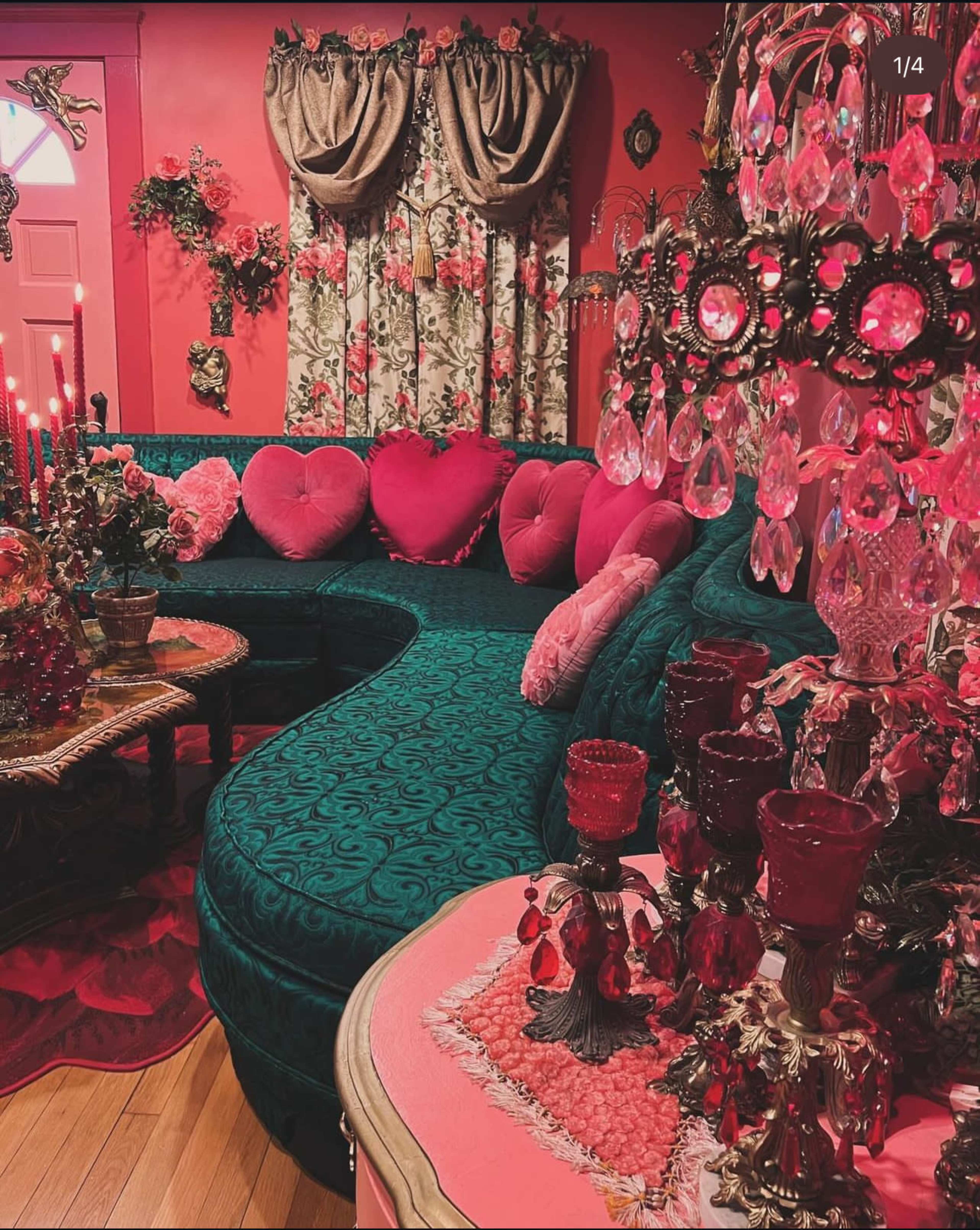 A brightly decorated living room features a teal sectional sofa adorned with pink heart-shaped pillows, alongside ornate candle holders and floral curtains.