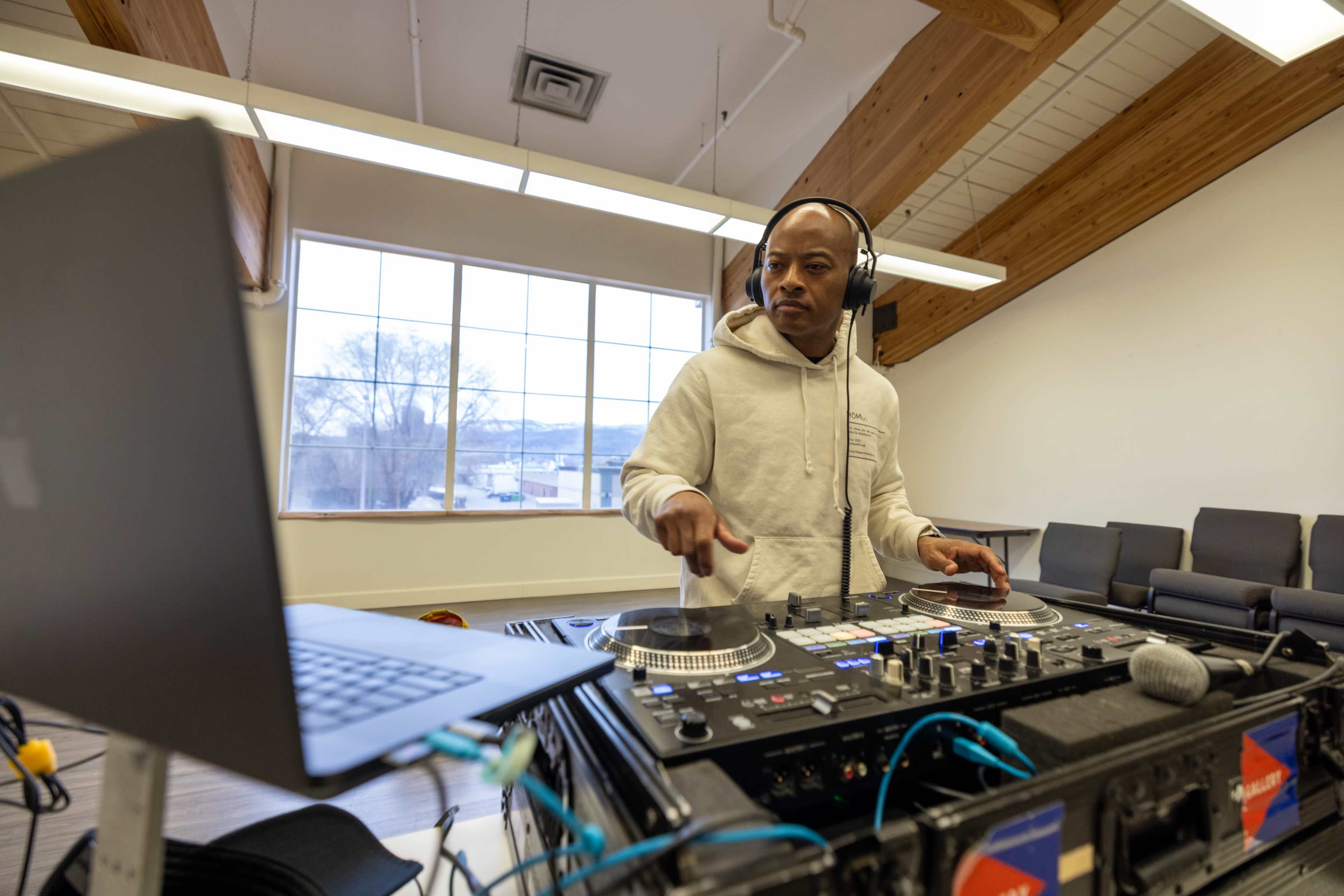A DJ wearing headphones mixes music on turntables in a spacious room with large windows and wooden beams.