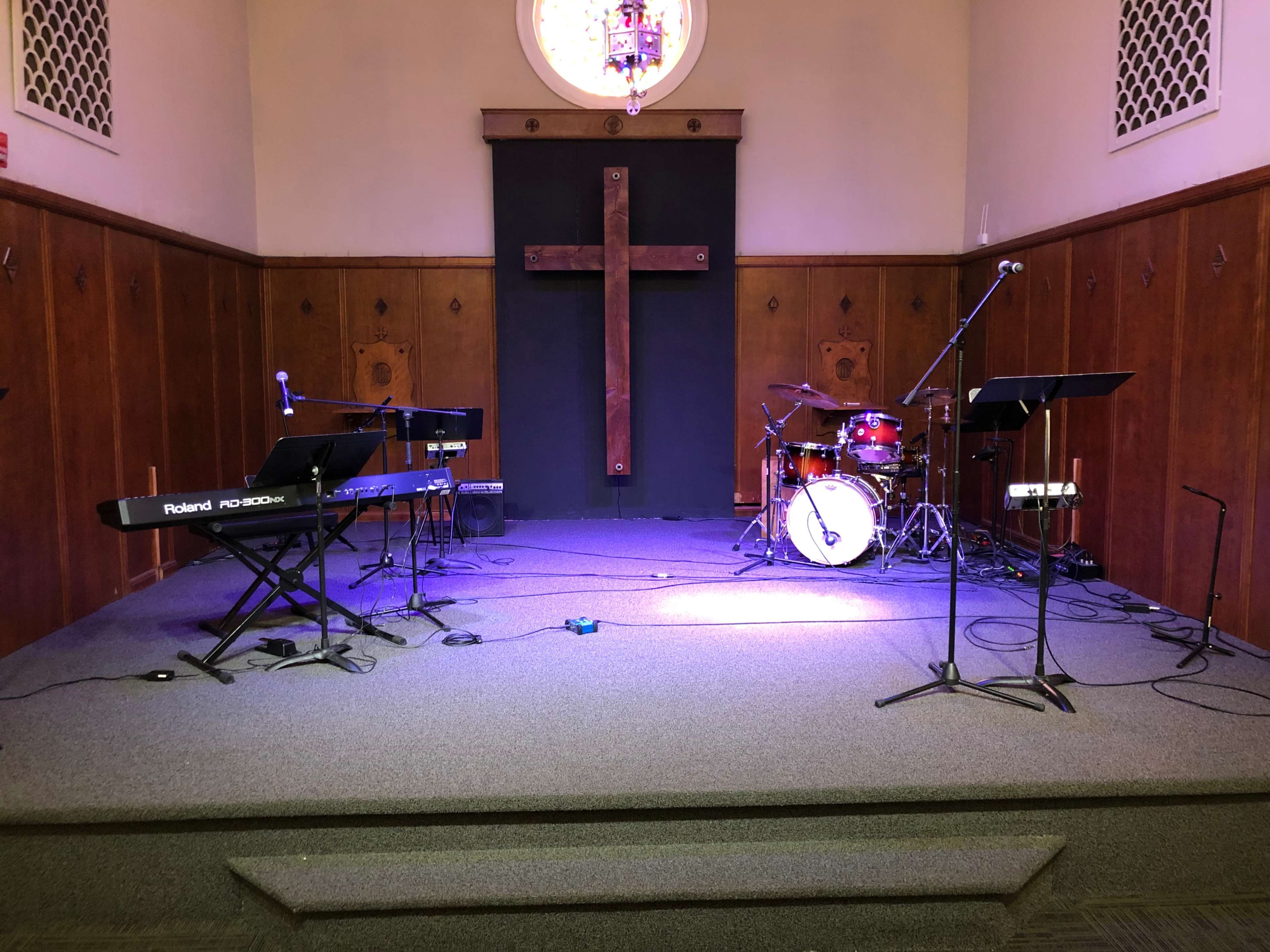 A stage setup in a church featuring musical instruments, including a keyboard, microphones, and a drum set, with a large wooden cross on the wall behind.