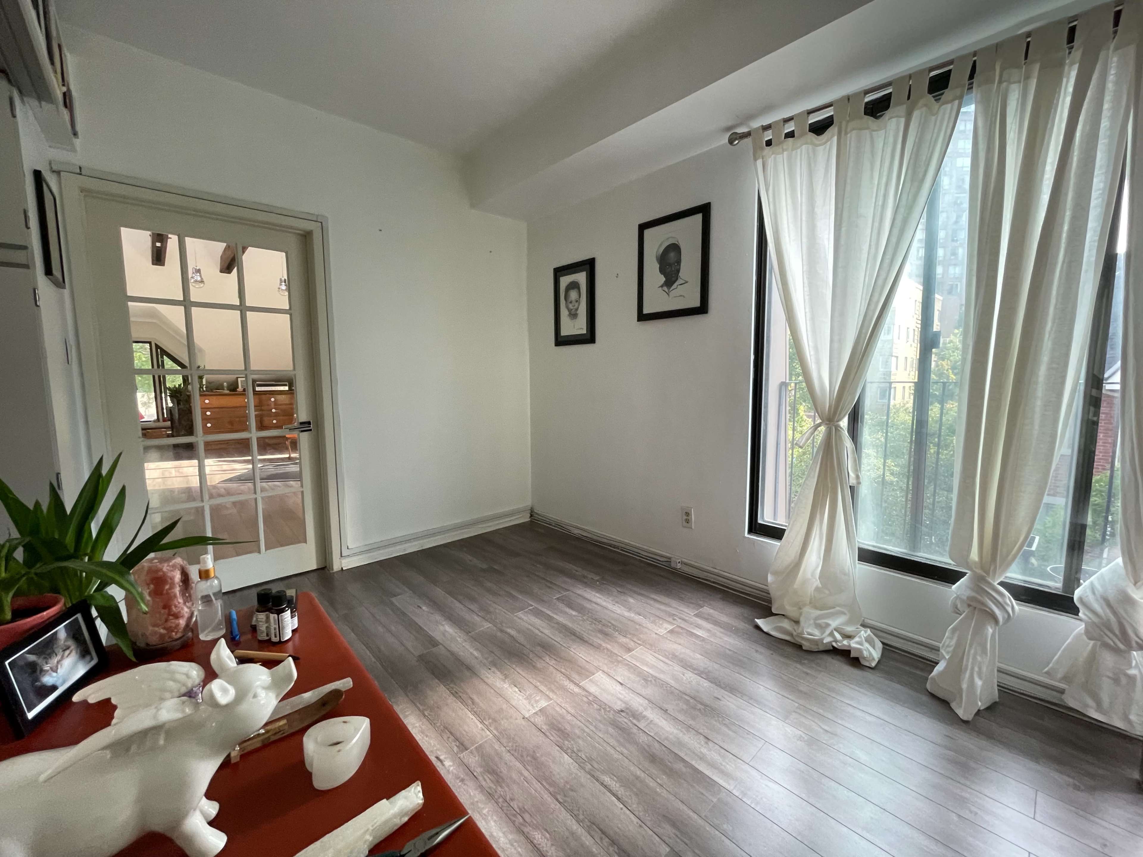 A bright room features large windows with sheer white curtains, hardwood flooring, and a small table with decorative items.