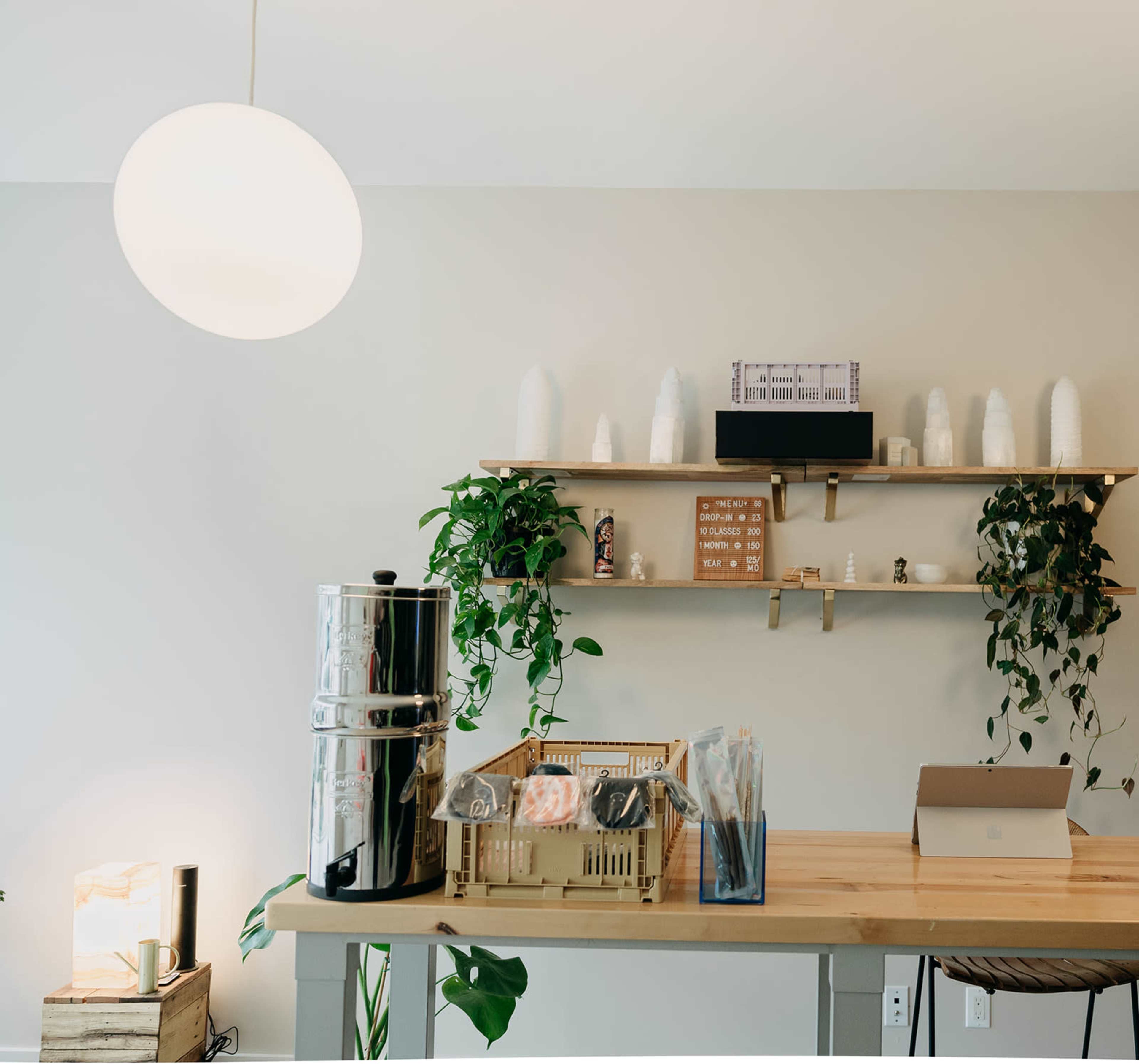 A well-lit room featuring a wooden table with a beverage dispenser and snacks, alongside shelves adorned with decorative items and plants.