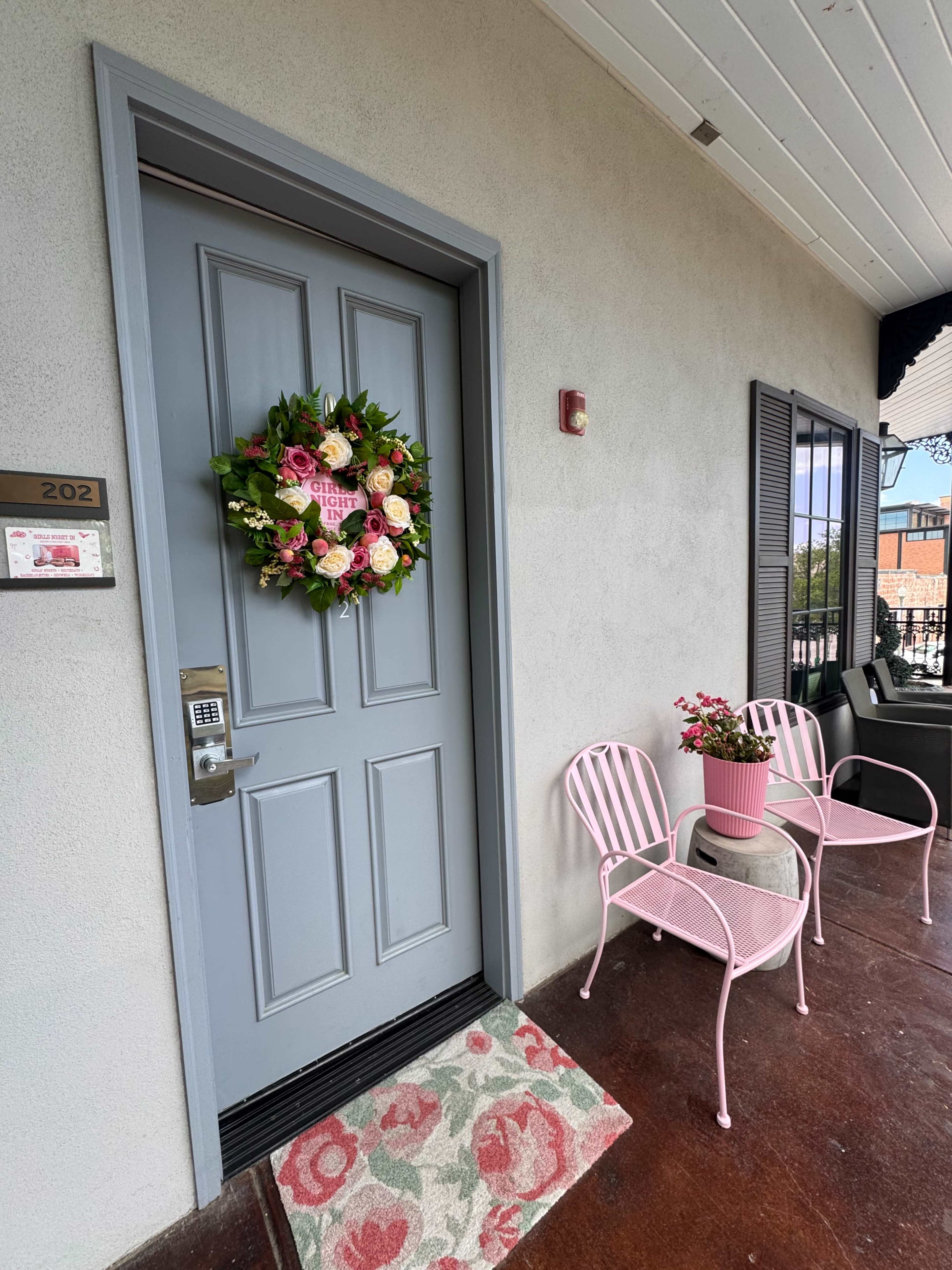 The image shows a gray apartment door adorned with a floral wreath, flanked by two pink metal chairs and a small table with a potted plant.