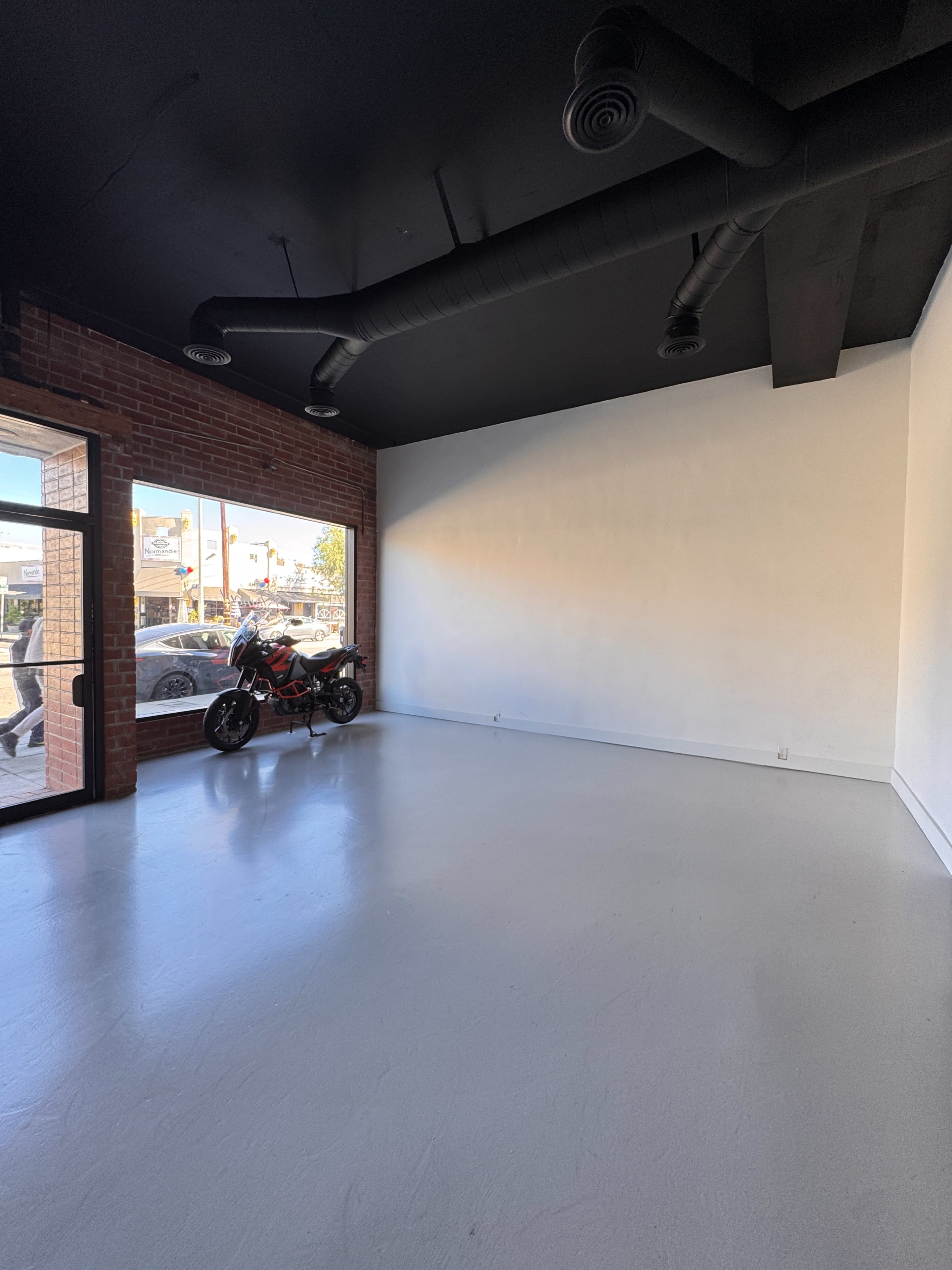 The image shows an empty interior space with a motorcycle and large windows providing natural light, alongside exposed brick and black ceiling vents.