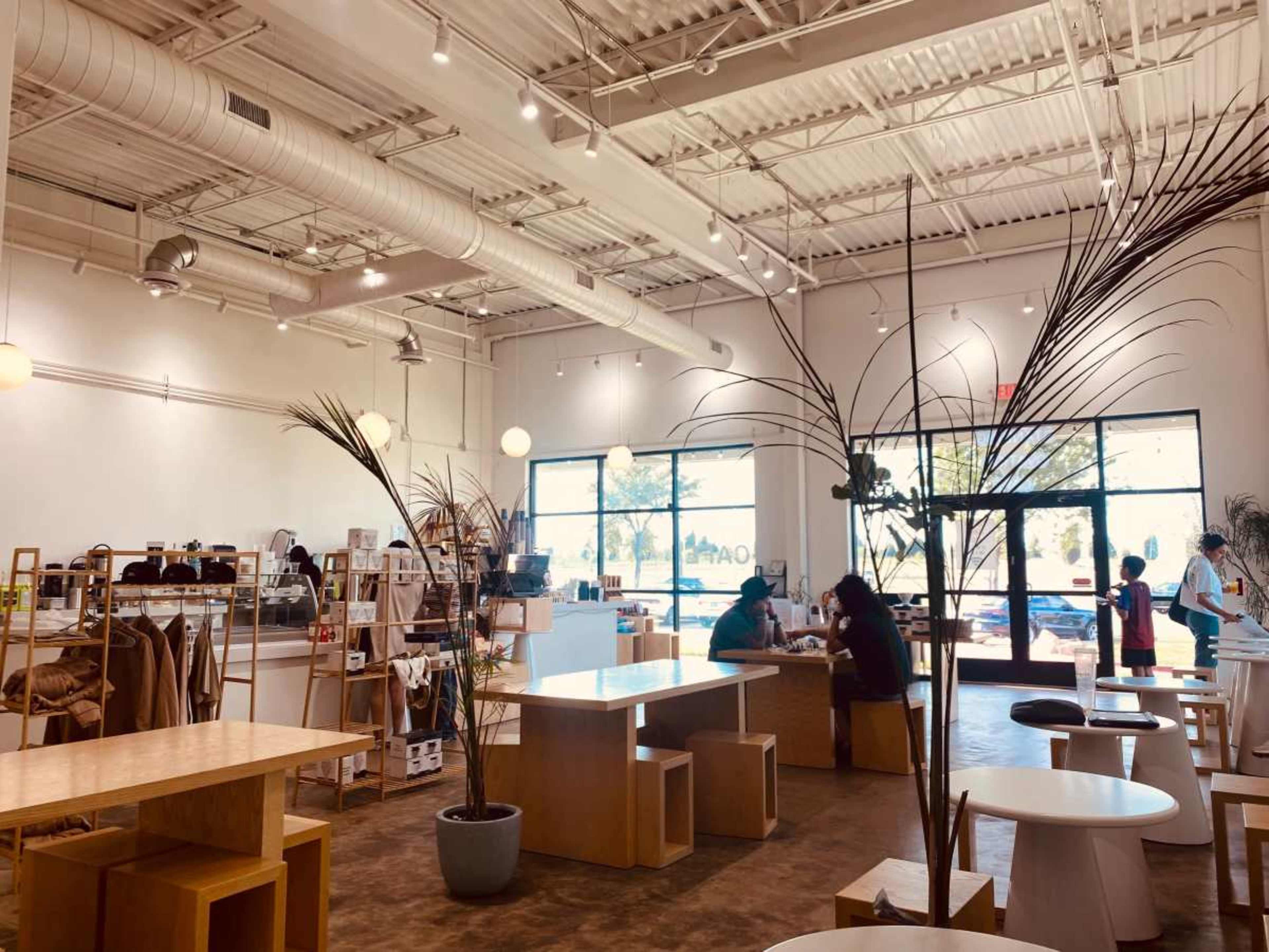 The image shows a modern cafe interior with wooden furniture, large windows letting in natural light, and customers seated at various tables.