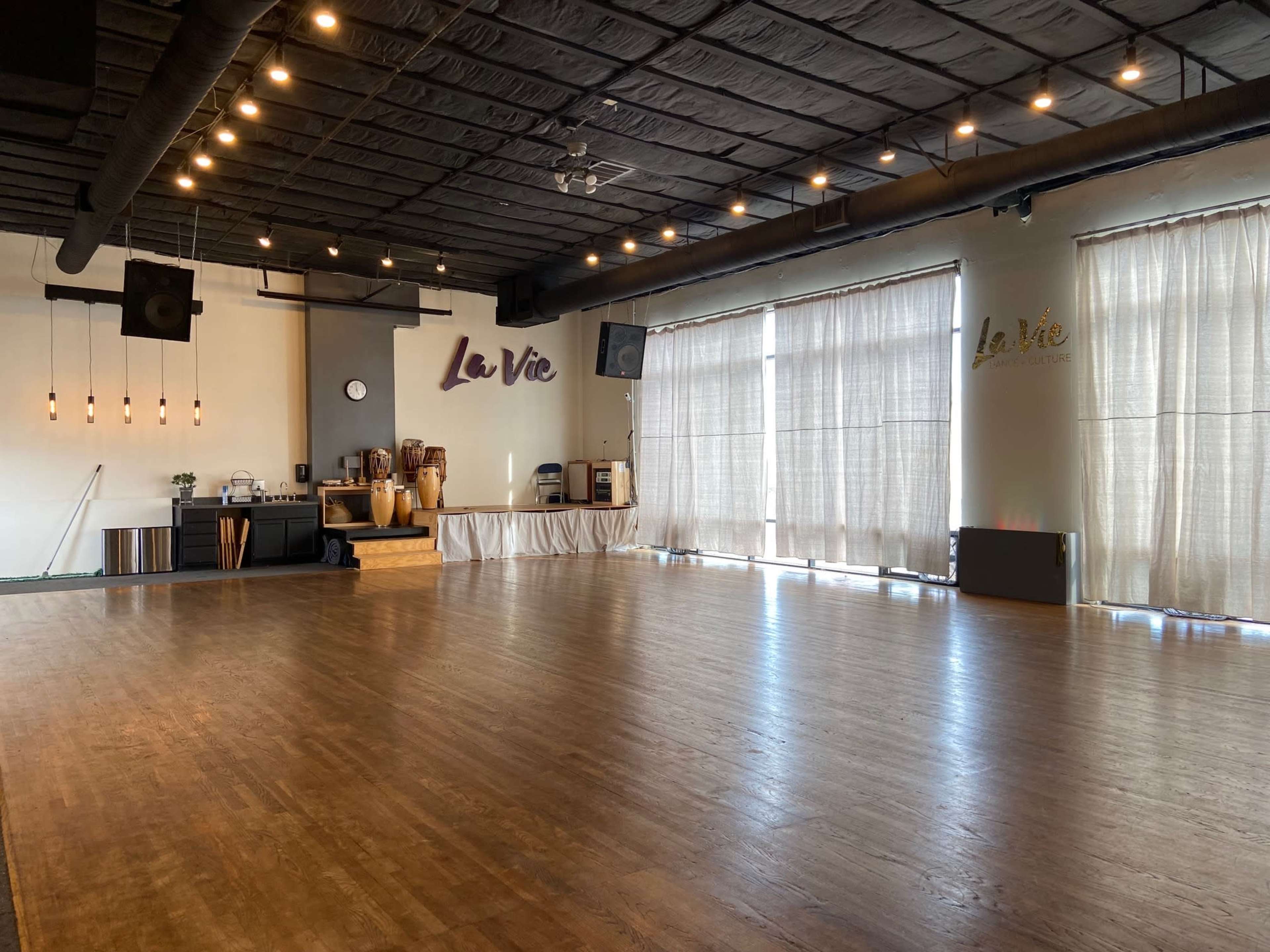 An empty dance studio with wooden flooring, large windows covered by sheer curtains, and a wall featuring the name "La Vie."