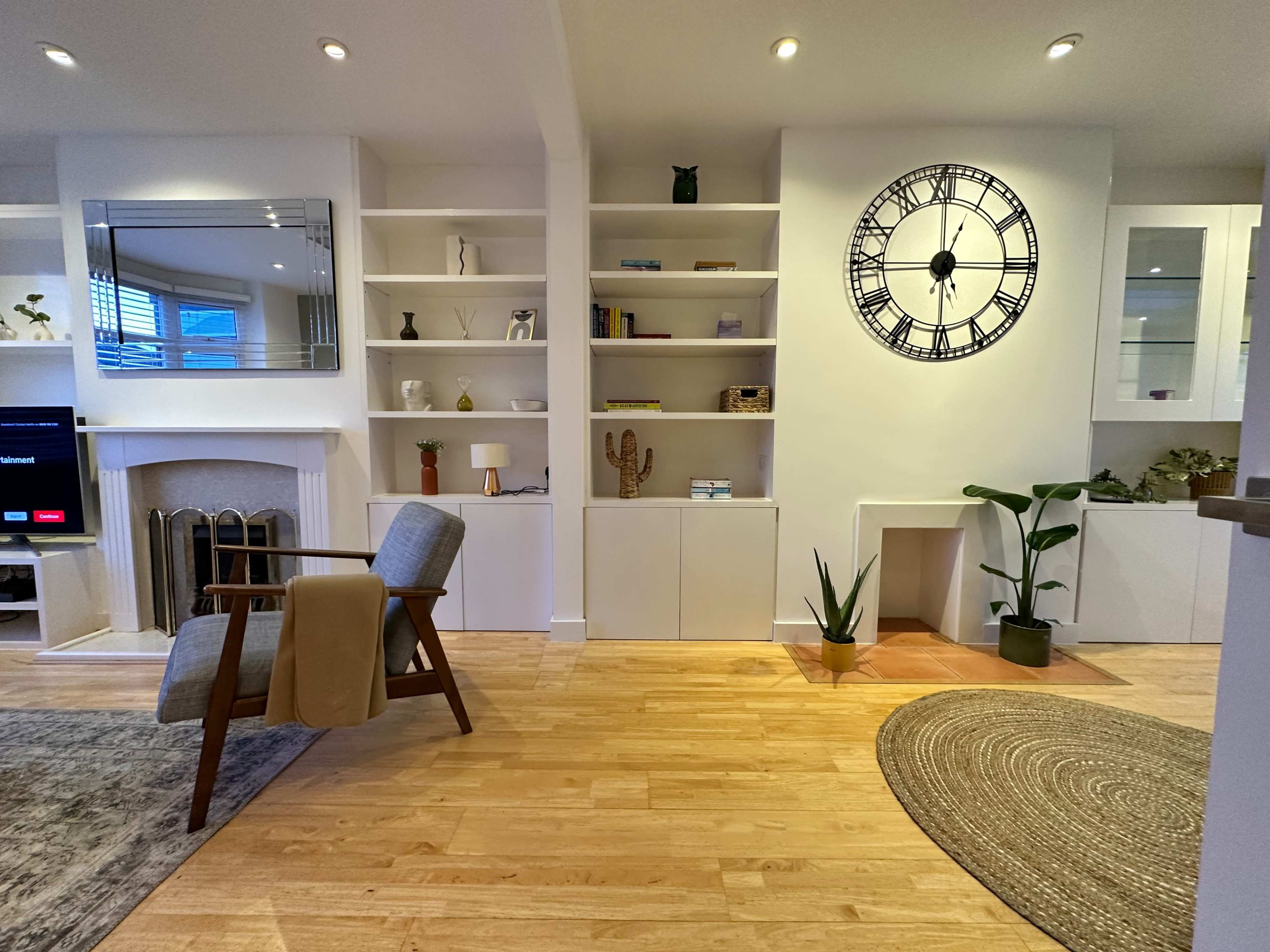 The living room features a comfortable chair, a wall clock, built-in shelves, and indoor plants, with wooden floors and a round rug.