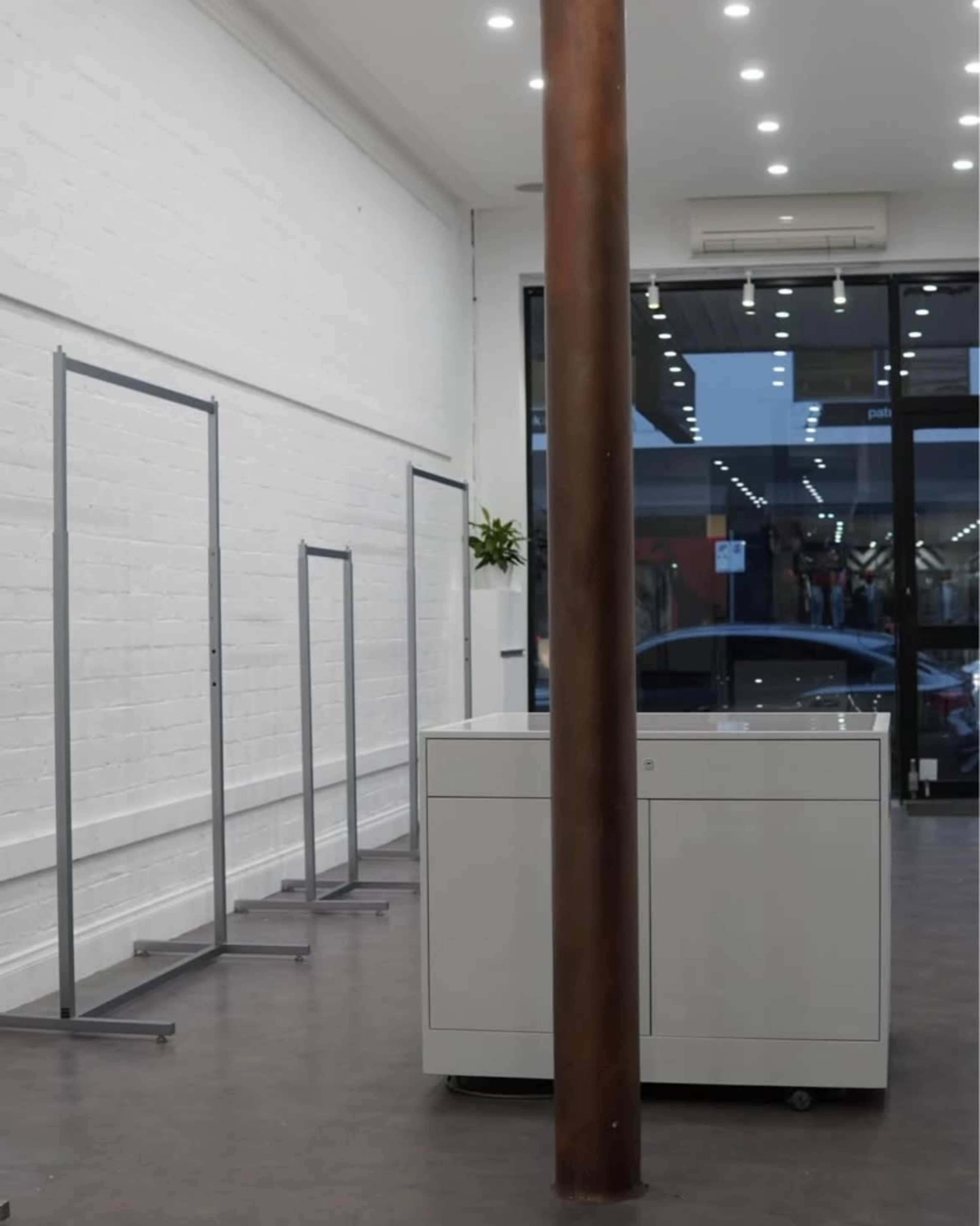 The image shows an interior space with several clothing racks along one wall and a white display table in the center, all set against a backdrop of large windows.