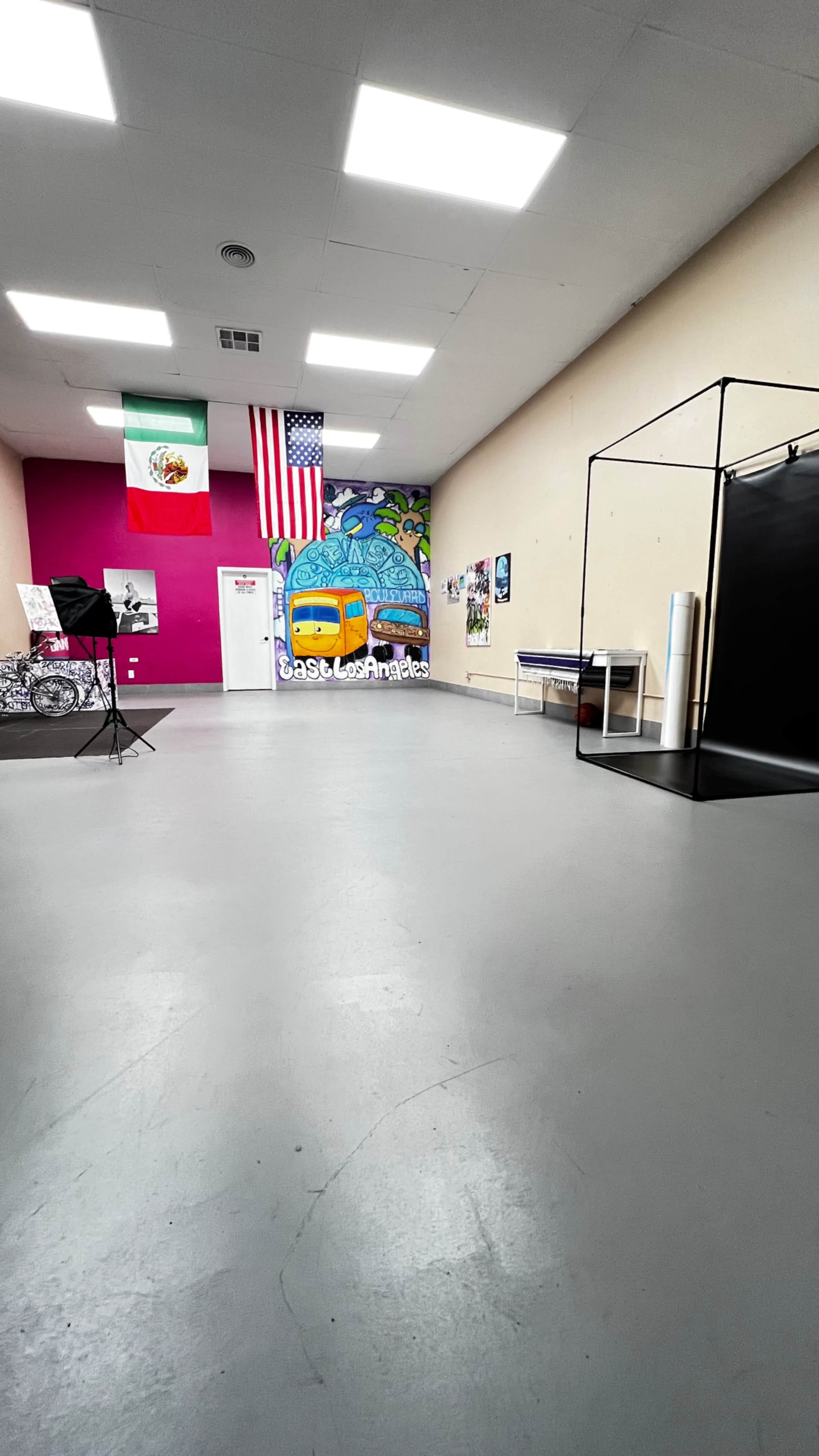 The image shows a spacious, well-lit room with gray flooring, a colorful wall featuring a mural, and national flags of Mexico and the United States displayed nearby.