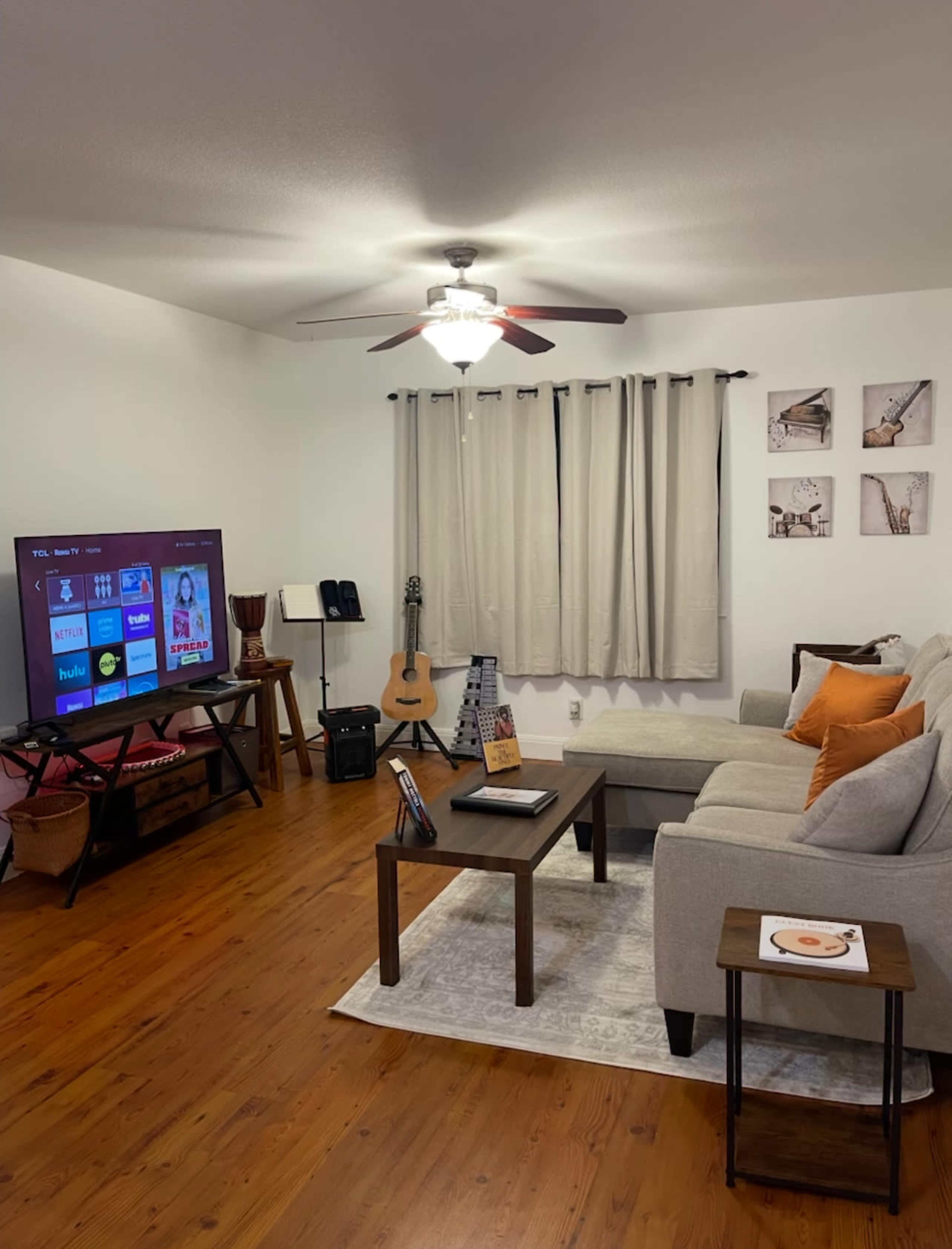 A cozy living room features a large television on a stand, a sectional sofa with orange pillows, a coffee table, and various musical instruments in the background.