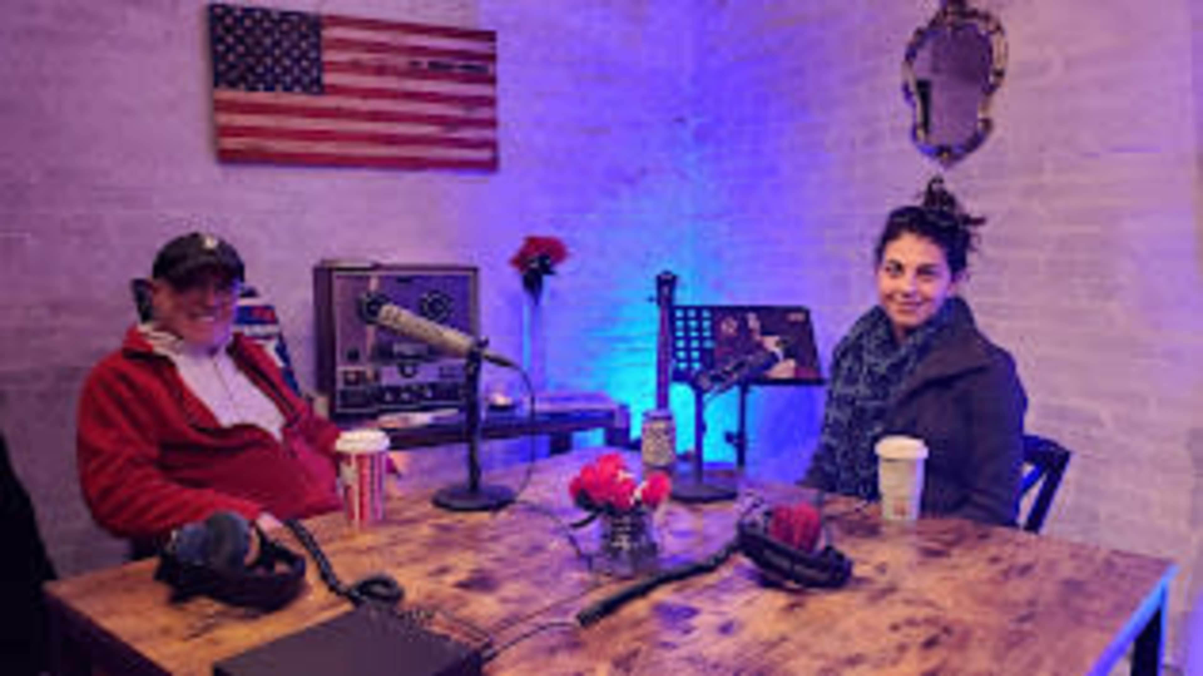 A man and a woman sit at a wooden table in a recording studio, with microphones, coffee cups, and an American flag on the wall.