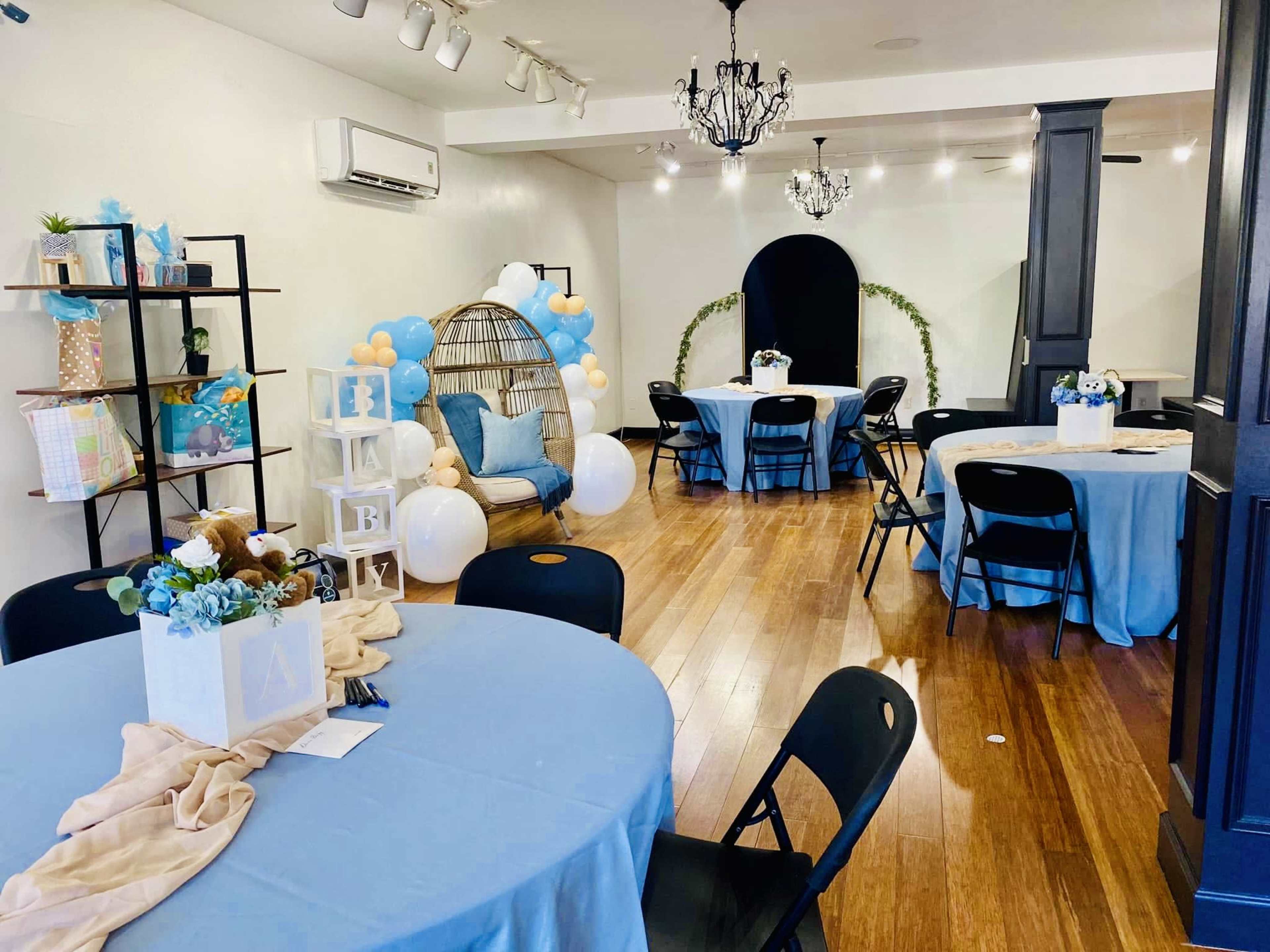 The image shows a decorated event space with round tables, a lounge area featuring a hanging chair, and various blue and white decorations.