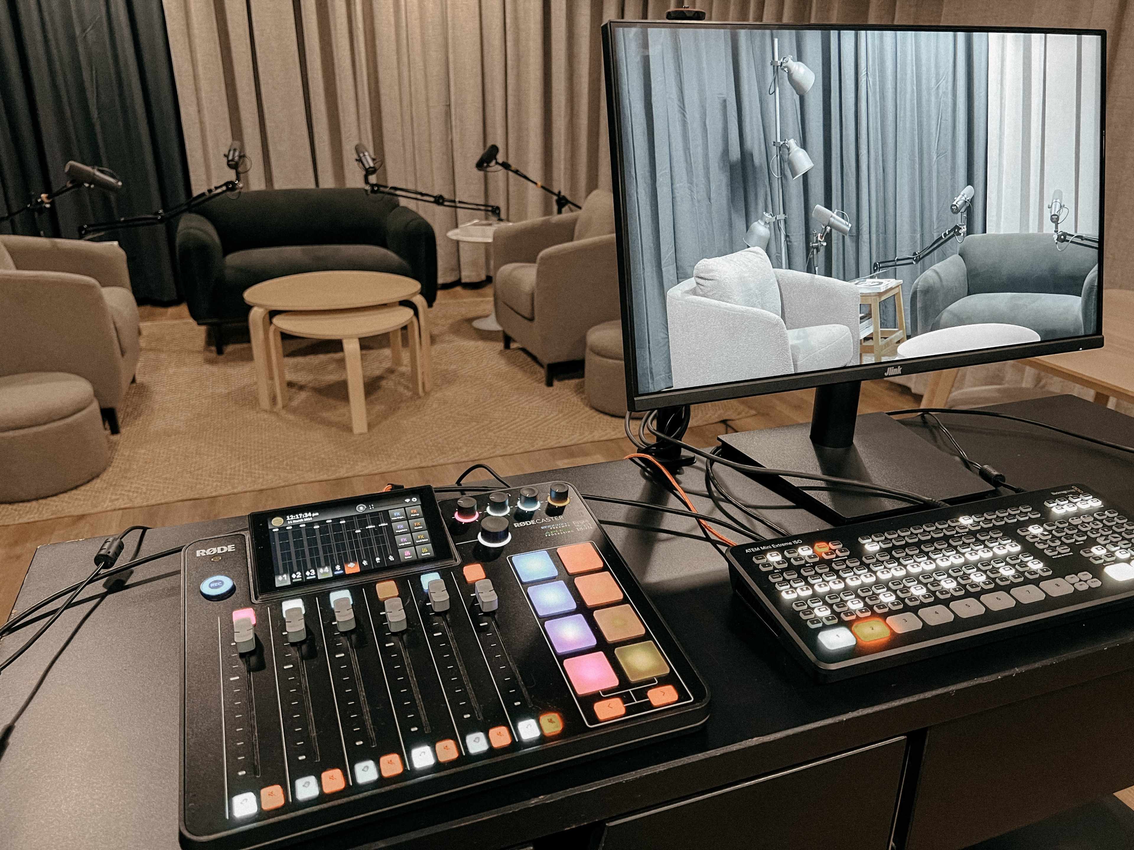 A video production studio is equipped with a sound mixer and keyboard, overlooking a seating area with microphones and a monitor displaying the scene.