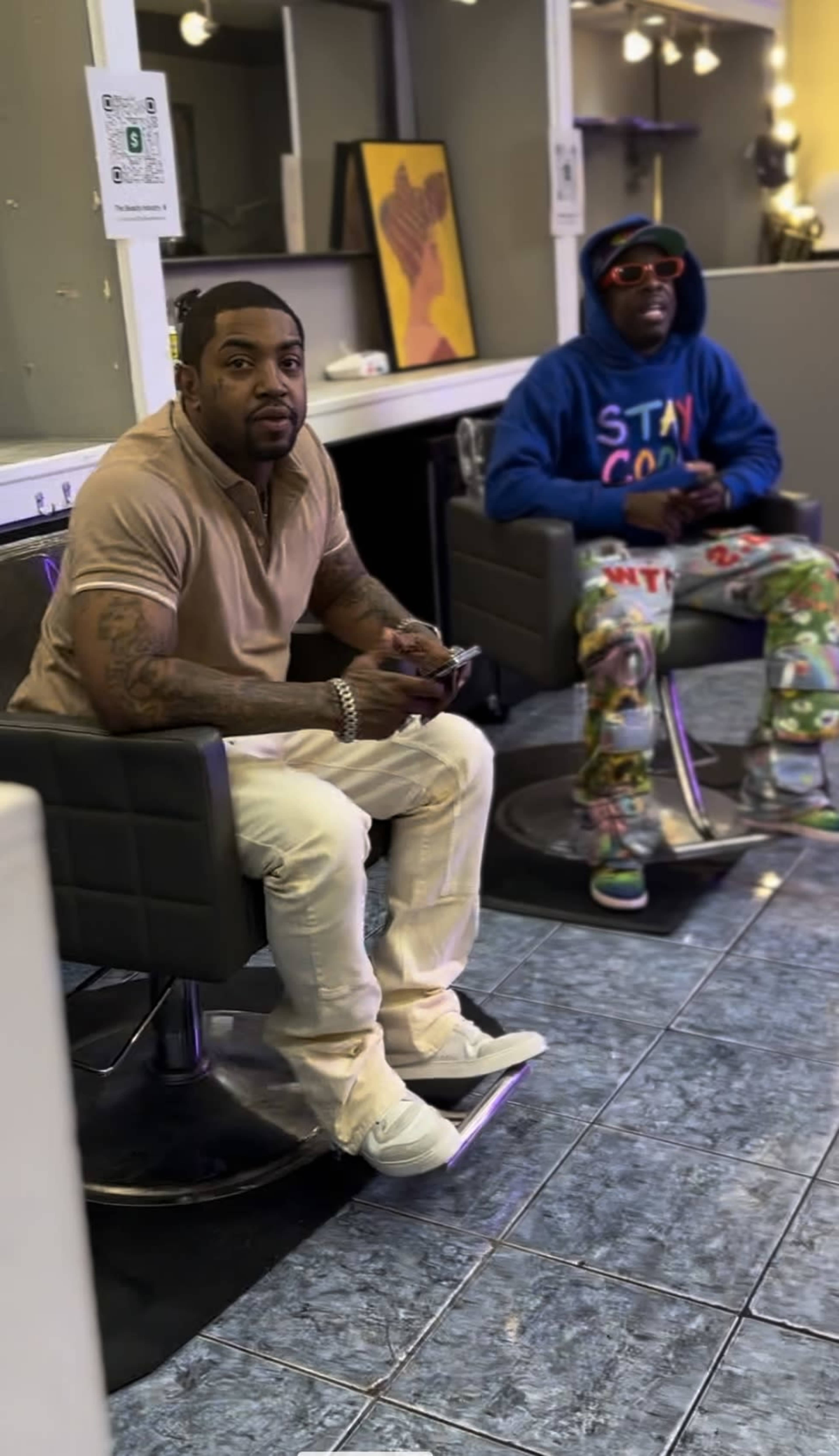 Two men are sitting in black chairs in a barbershop, one wearing a beige shirt and the other in a colorful hoodie and printed pants.