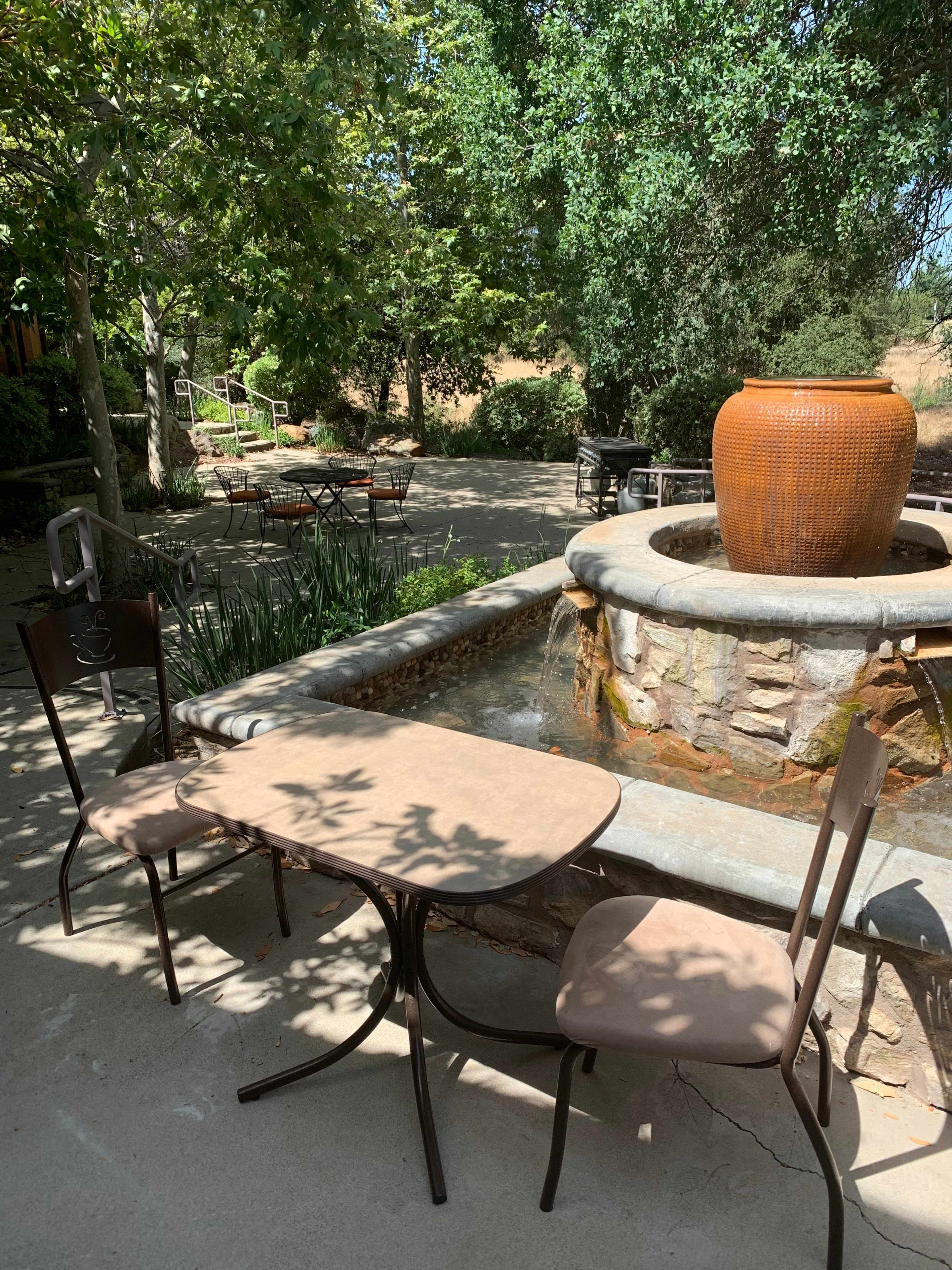 A small outdoor seating area features two chairs and a table beside a stone fountain surrounded by greenery.