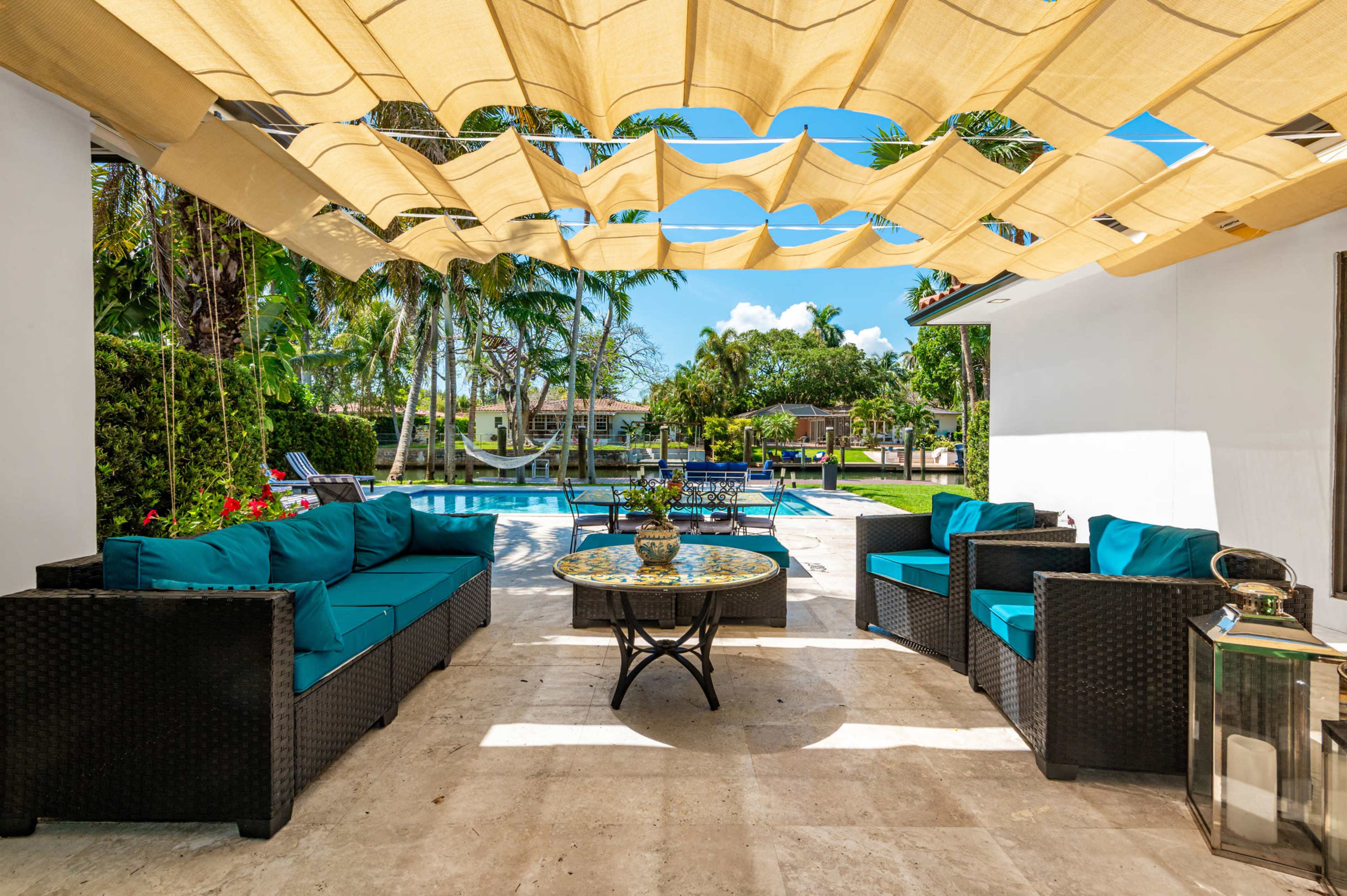 A seating area with blue cushions is set under a fabric canopy, overlooking a pool surrounded by tropical plants.