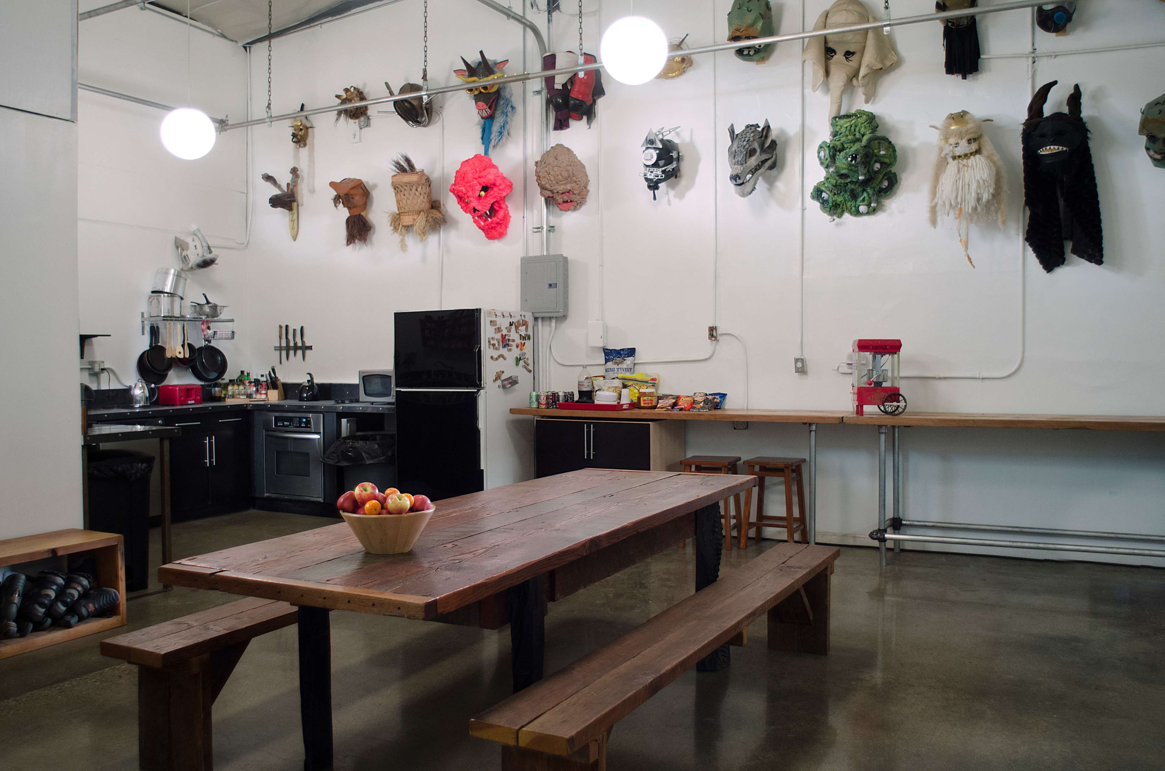 The room features a long wooden table with a bowl of apples, surrounded by kitchen appliances and masks displayed on the walls.