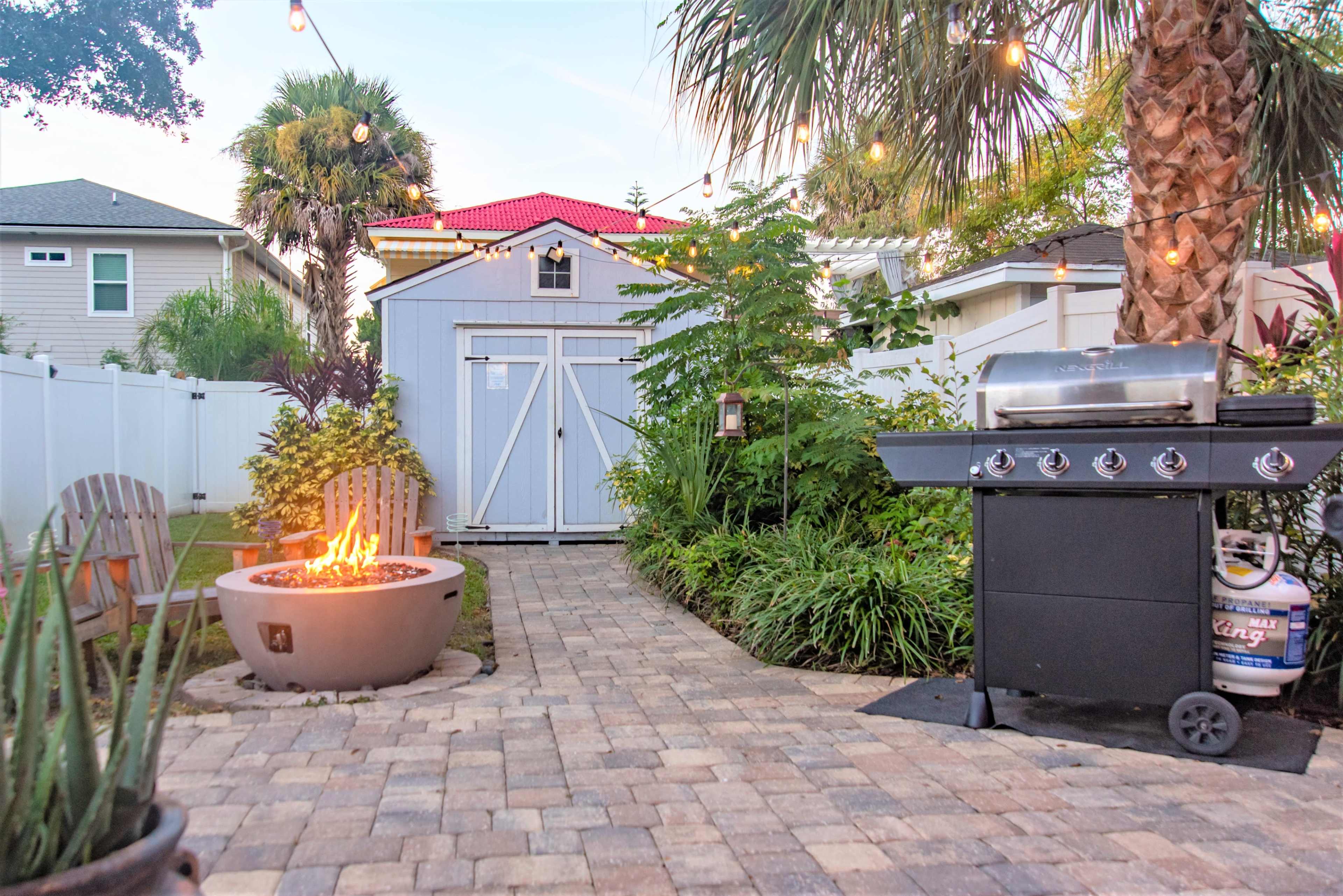 A backyard with a stone pathway leading to a fire pit and a barbecue grill, surrounded by lush greenery and a small shed.