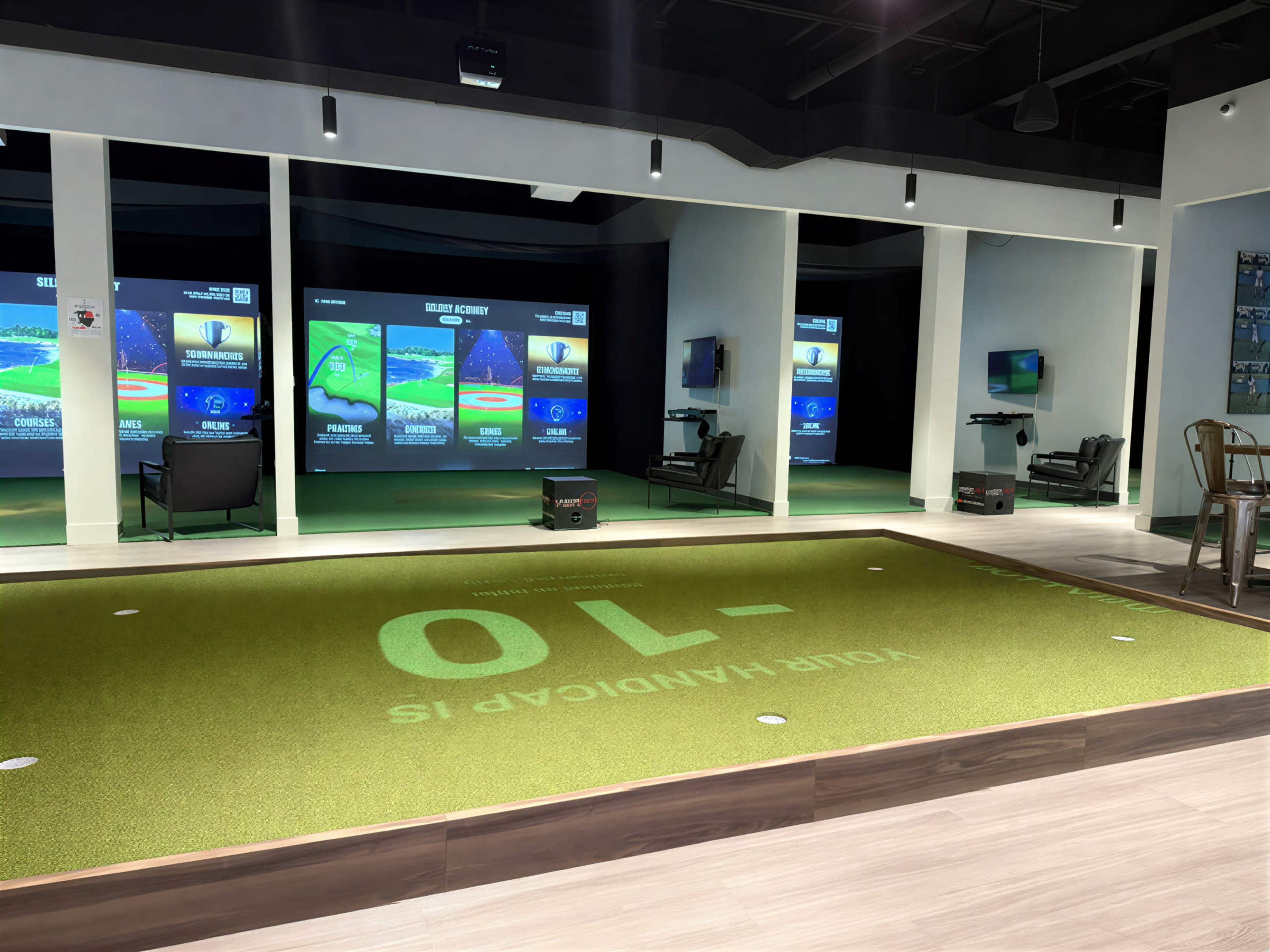 The image shows an indoor golf simulation facility featuring a putting green in the foreground and multiple large screens displaying golf course graphics in the background.