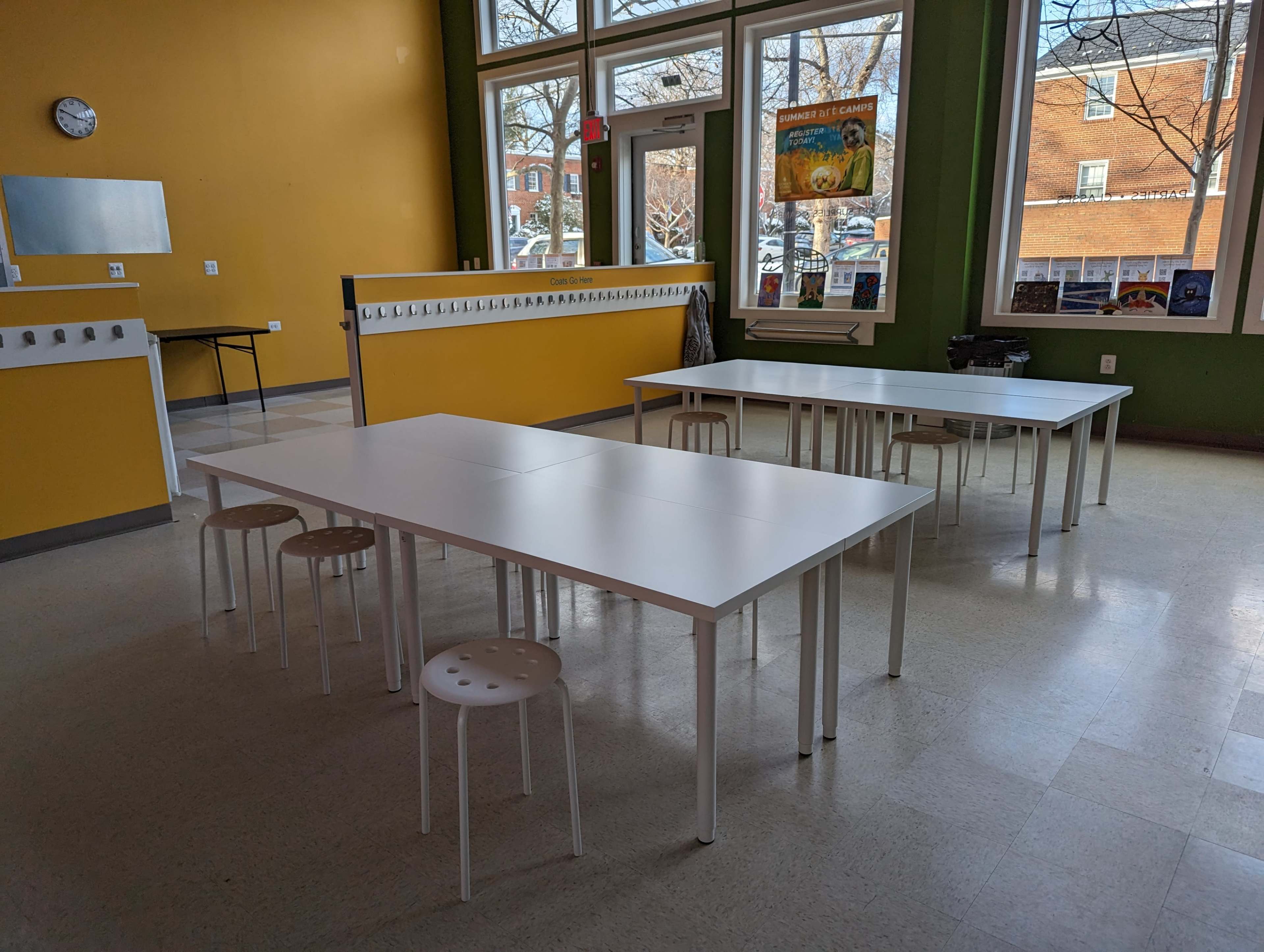 A classroom with several white tables and stools arranged in a spacious, well-lit area with large windows.