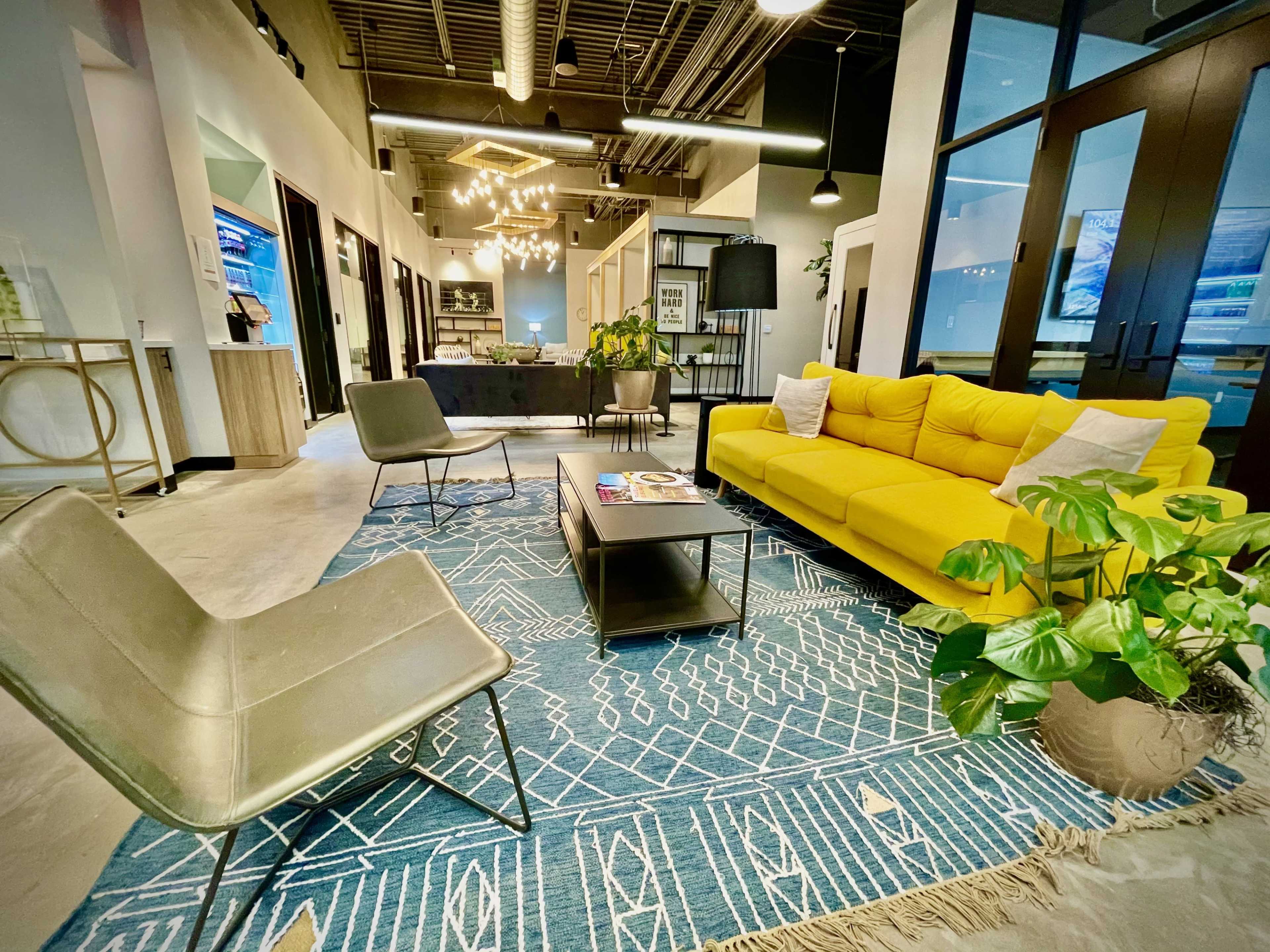 A modern office lounge area featuring a bright yellow sofa, two gray chairs, a coffee table, and a decorative plant on a blue patterned rug.