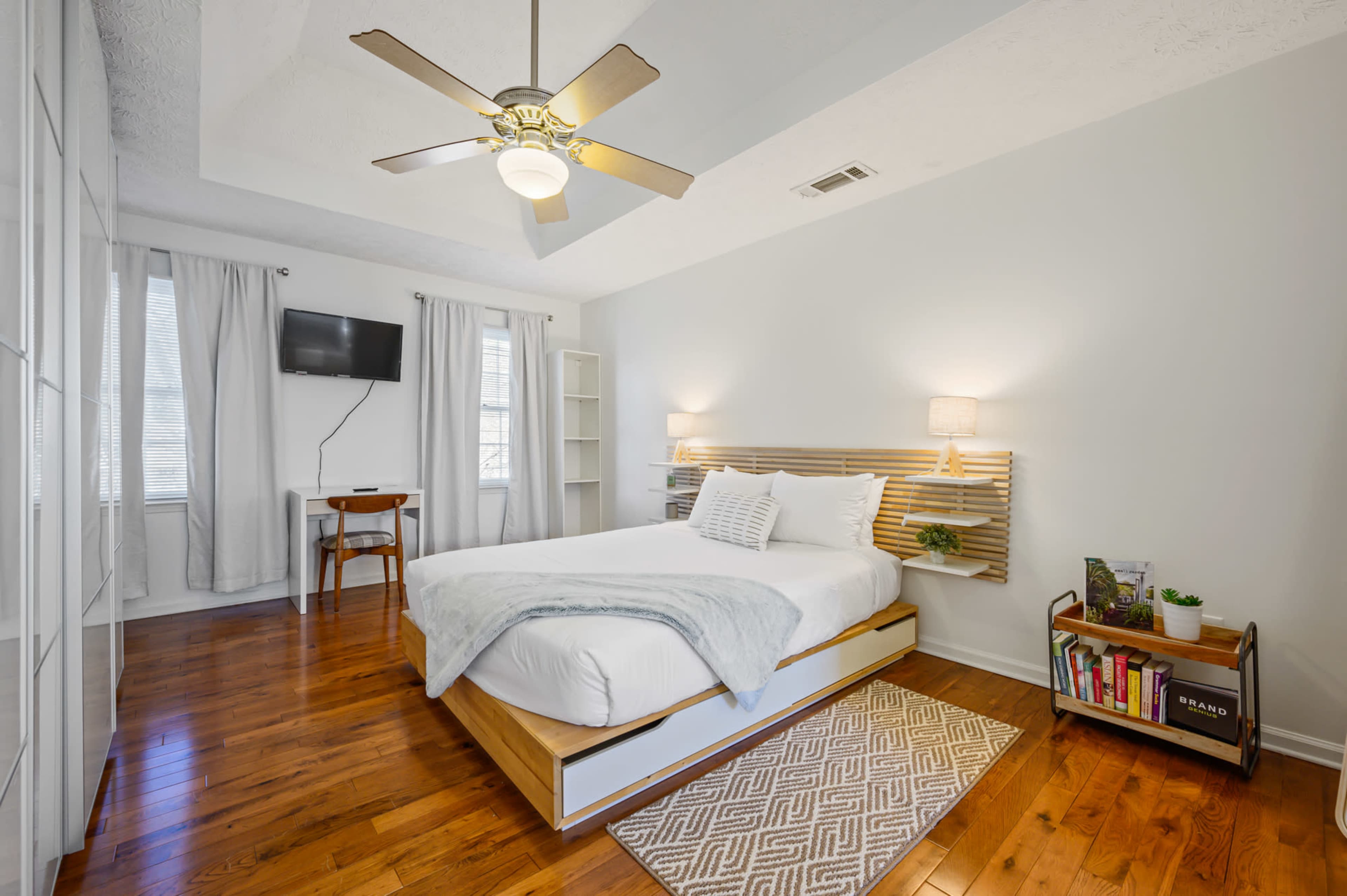 The image shows a well-lit bedroom featuring a queen-sized bed with a wooden headboard, a ceiling fan, a small desk, and a TV mounted on the wall.