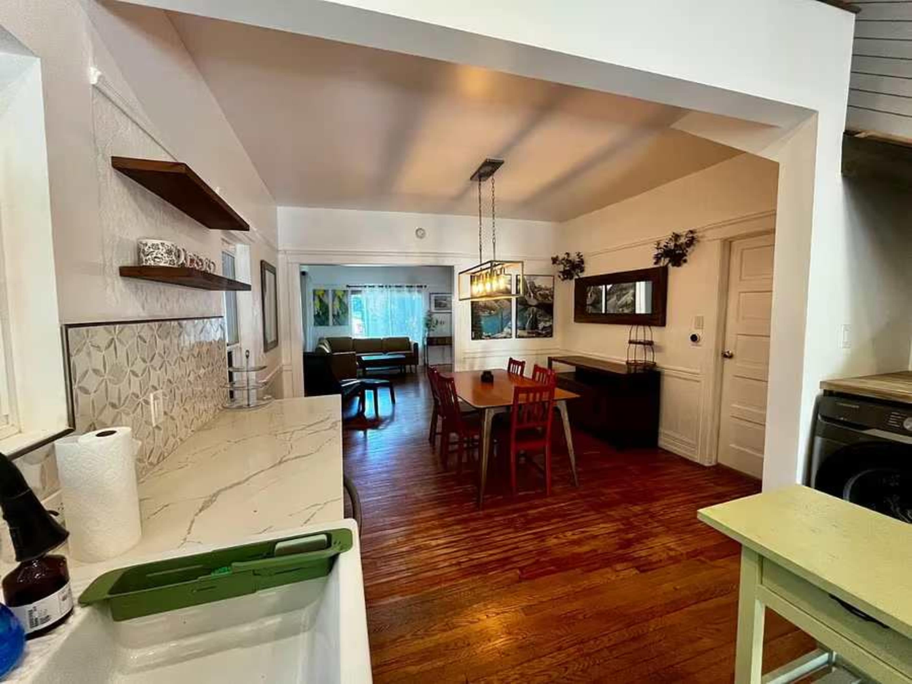 The image shows a well-lit kitchen open to a dining area, featuring wooden floors, a dining table with red chairs, and decor on the walls.