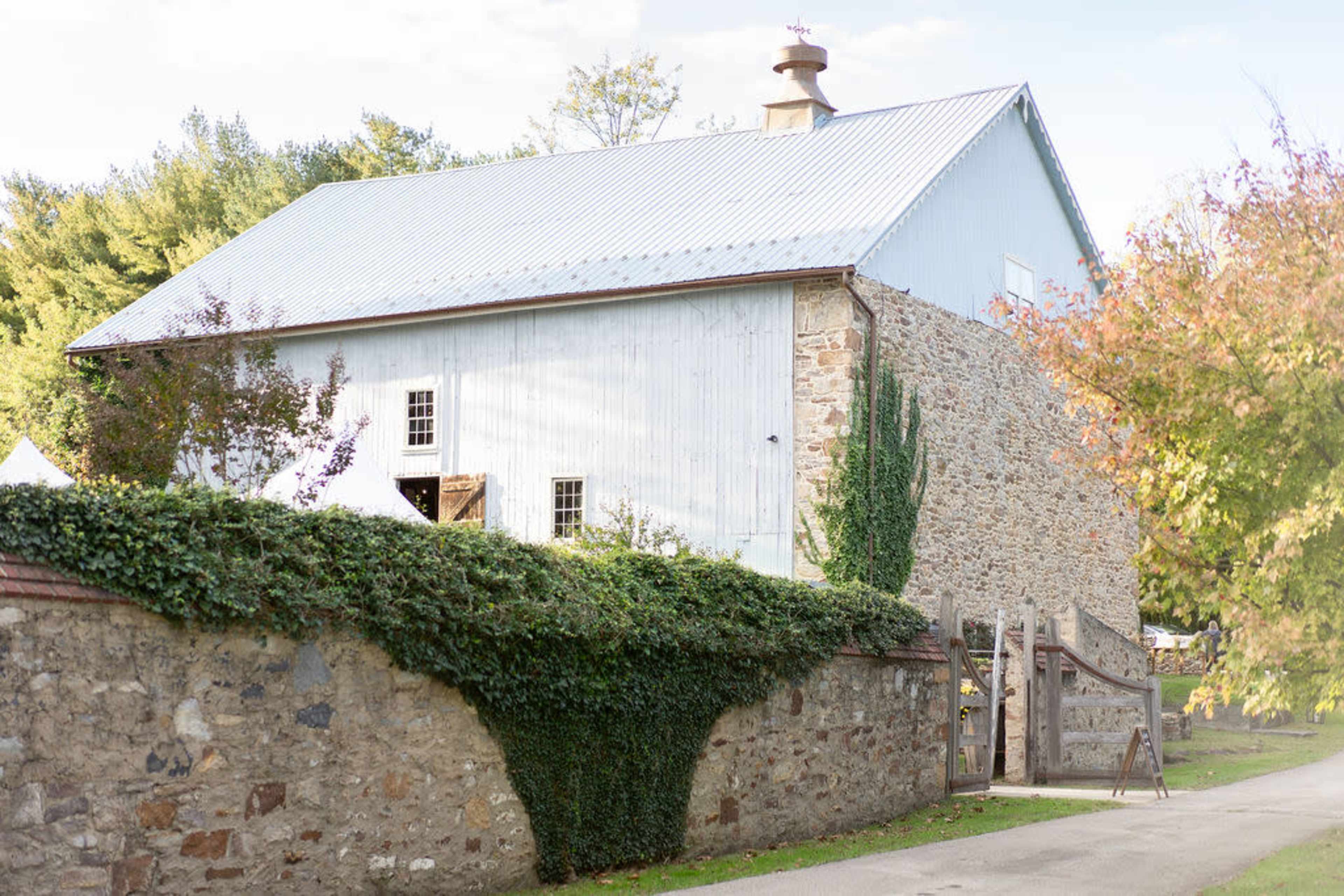 A stone barn with a metal roof sits beside a gravel pathway, surrounded by greenery and a stone wall.