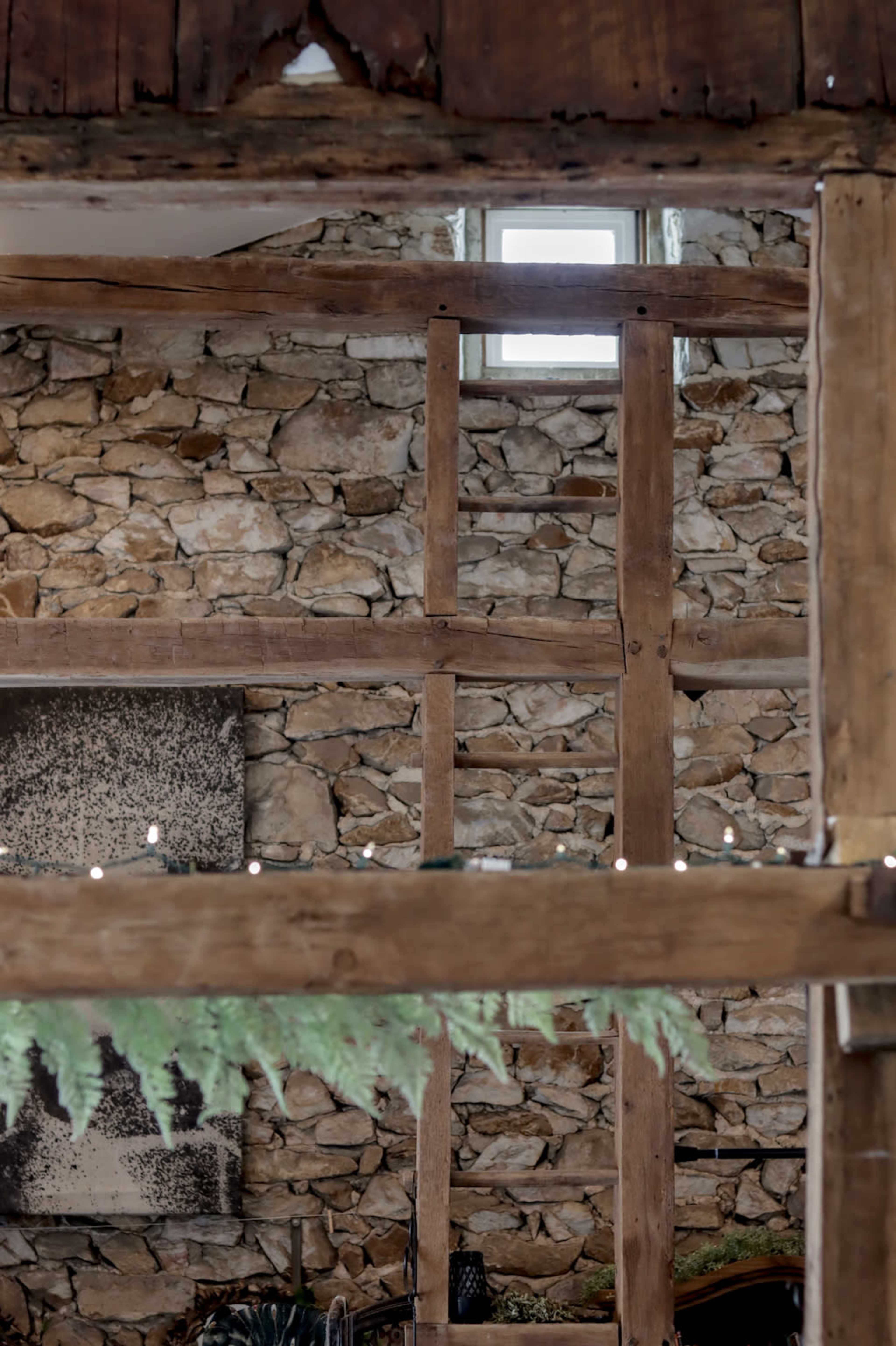 The image shows a rustic interior space with a stone wall, wooden beams, and a small window above.