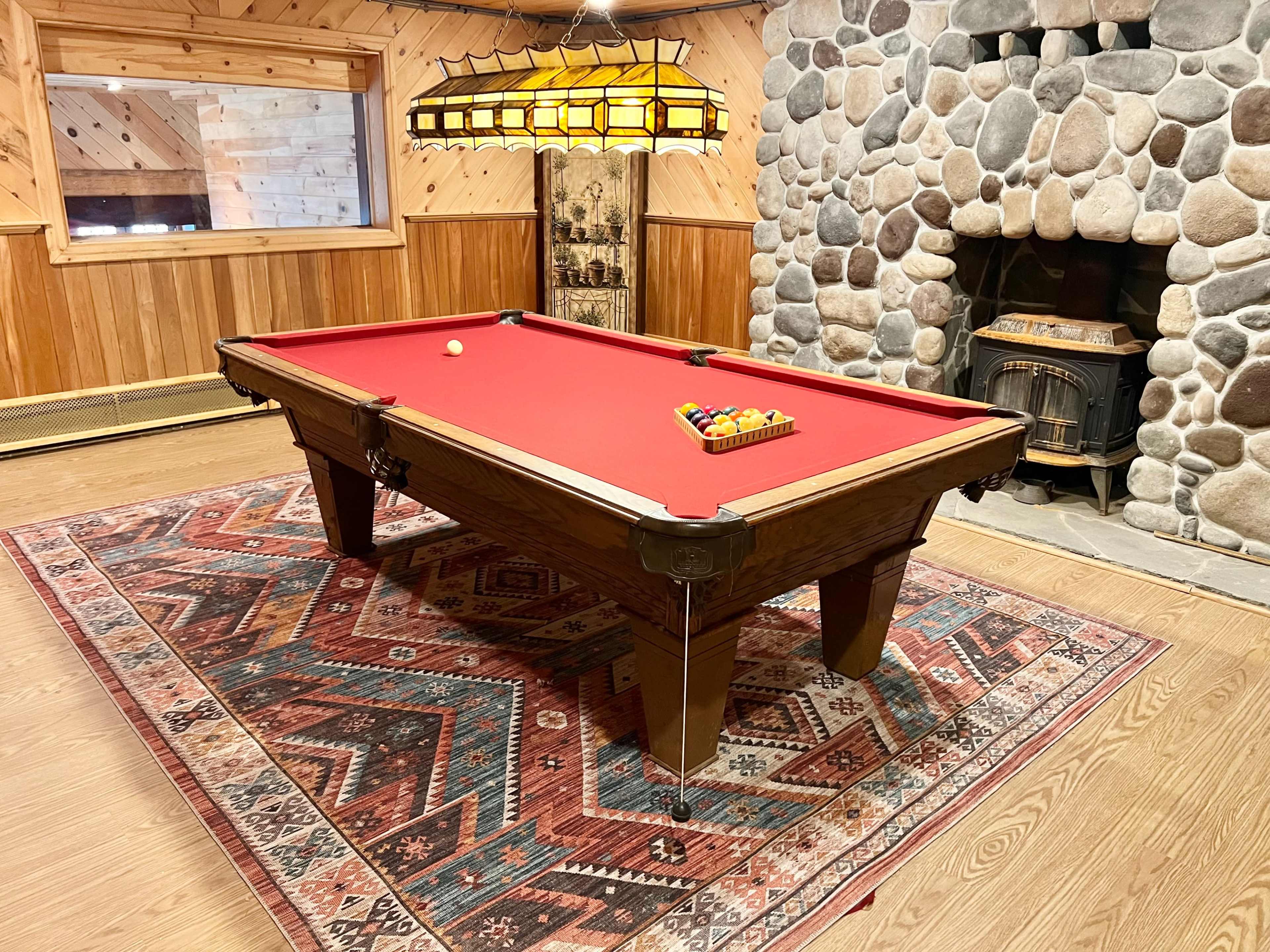 A red felt pool table with a set of billiard balls arranged on it, placed in a cozy room featuring a stone fireplace and wooden walls.