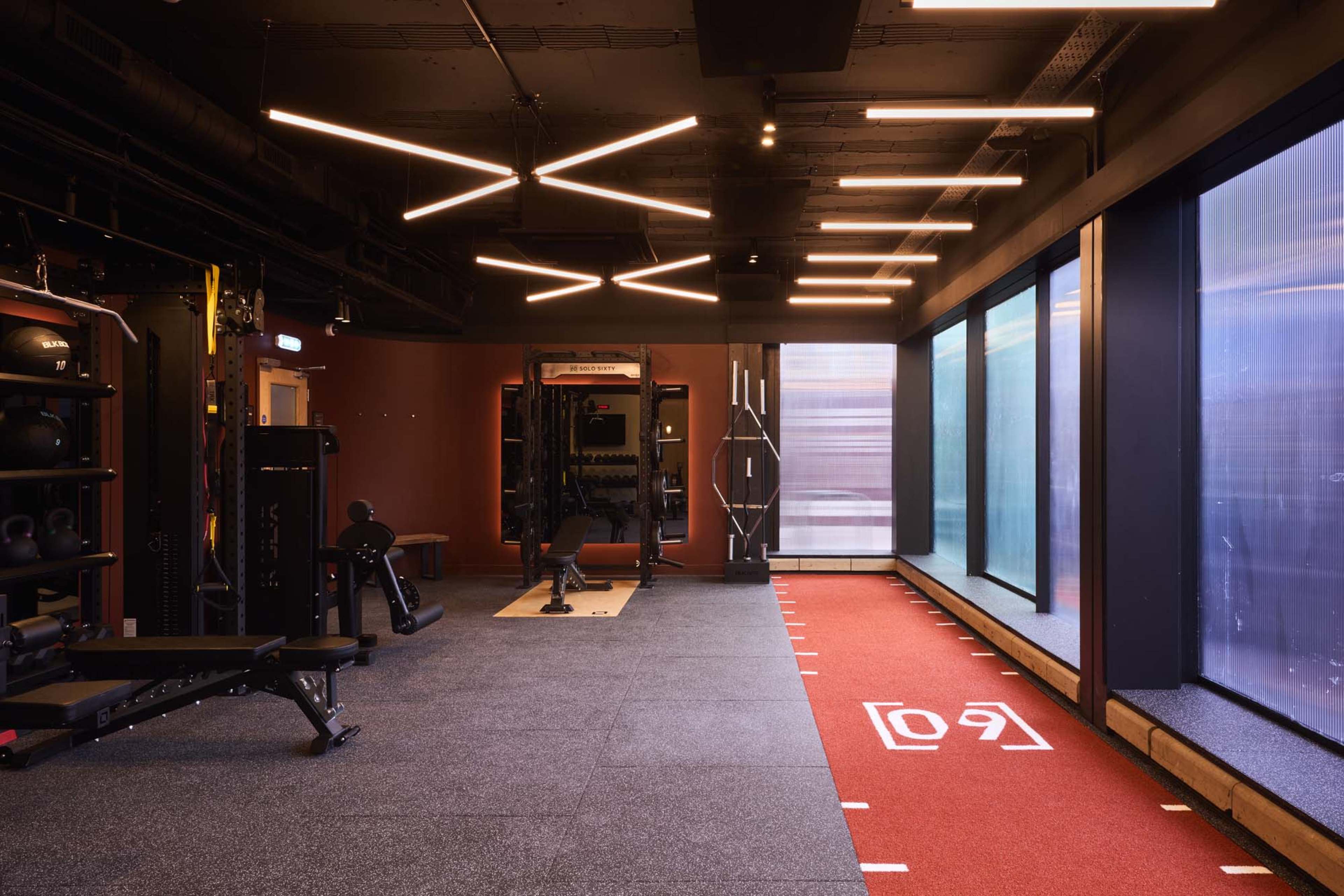 The image shows a modern gym interior featuring exercise equipment, dark flooring, and large windows along one wall.