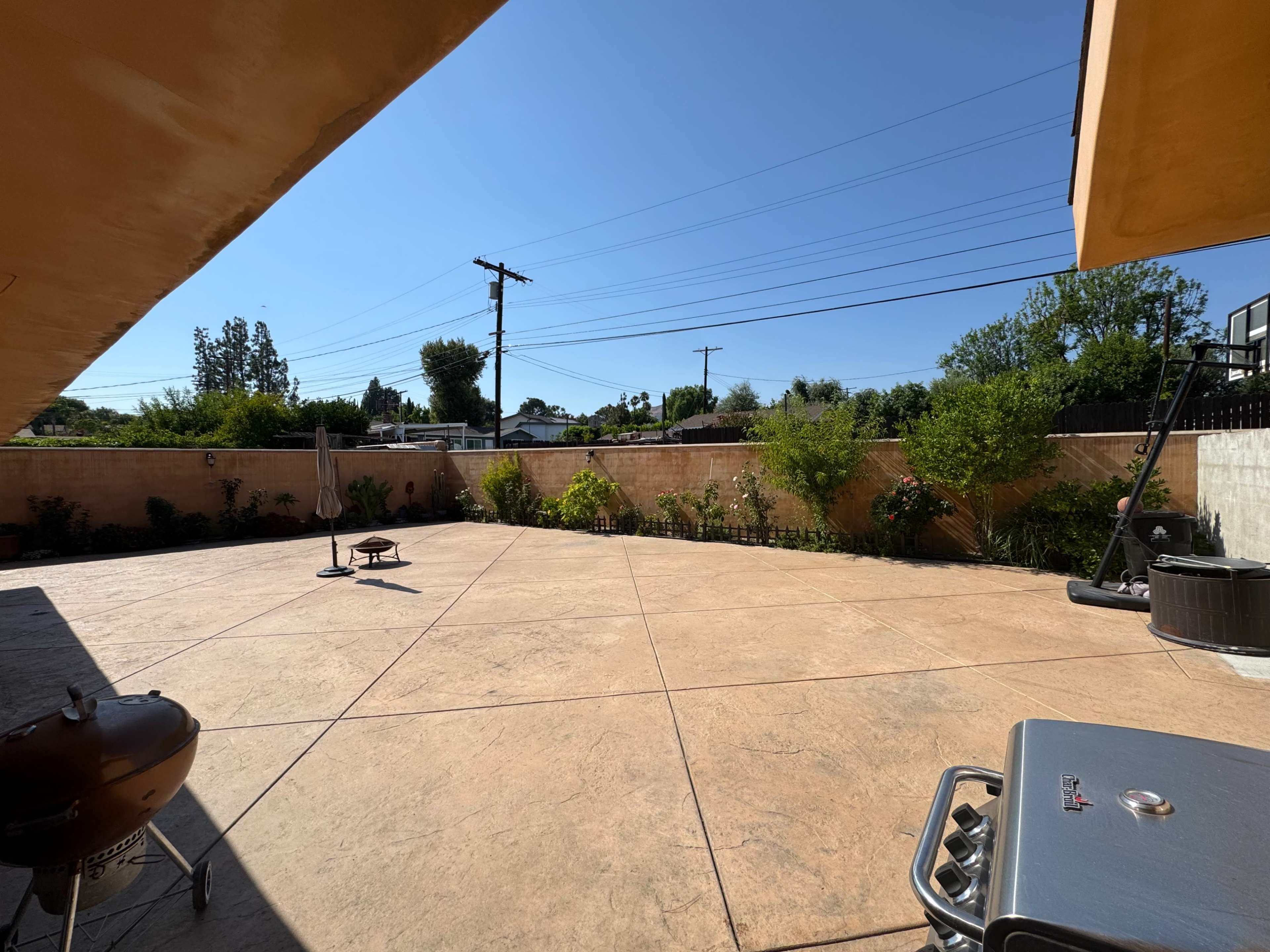 A spacious outdoor patio with a concrete surface, surrounded by plants and trees, features a grill and a small fire pit.