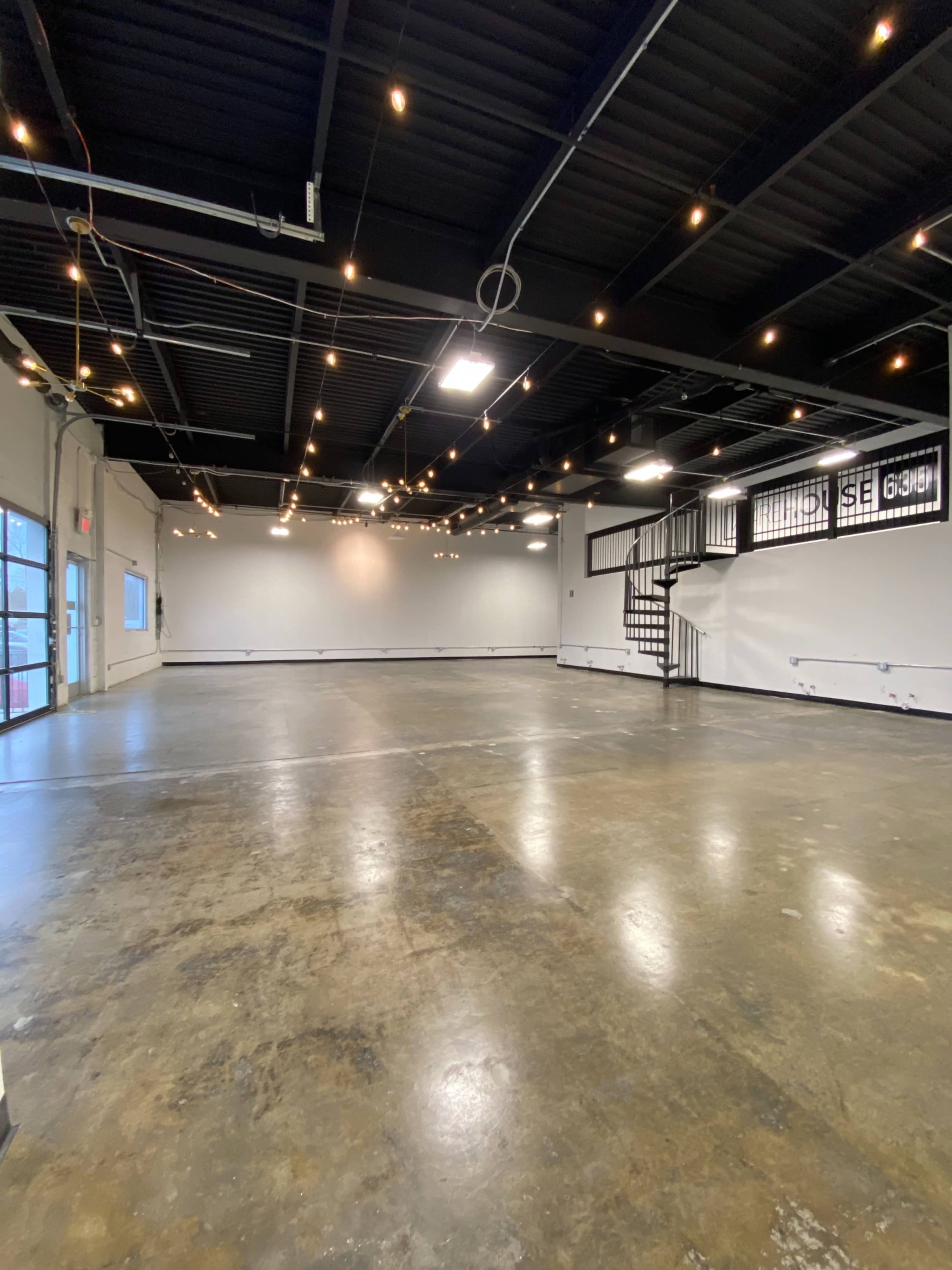 An empty industrial-style room with a polished concrete floor, string lights overhead, and a spiral staircase in the corner.