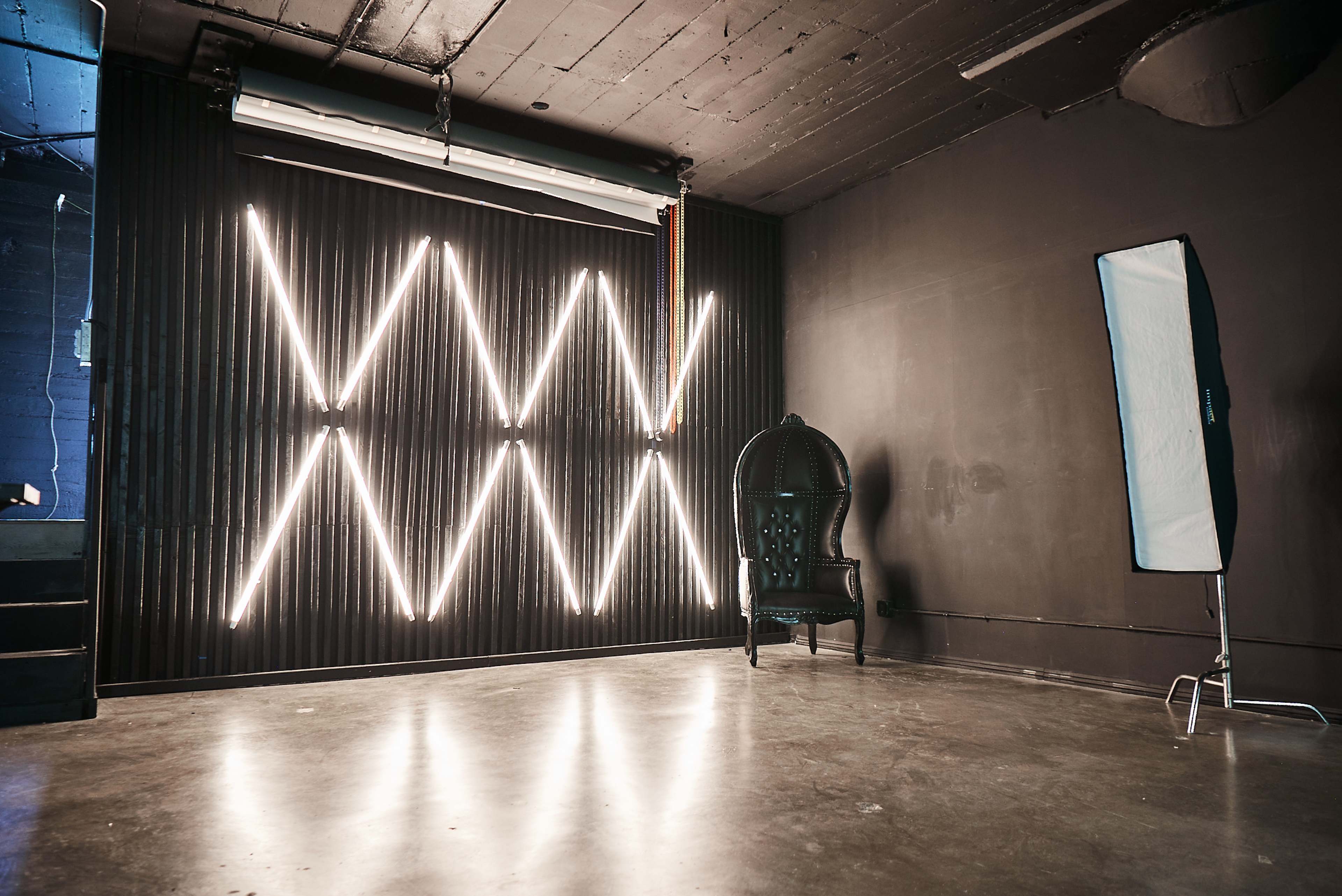 The image features a dimly lit room with a black wall illuminated by a geometric pattern of bright lights, and a vintage chair positioned beside a softbox lighting setup.