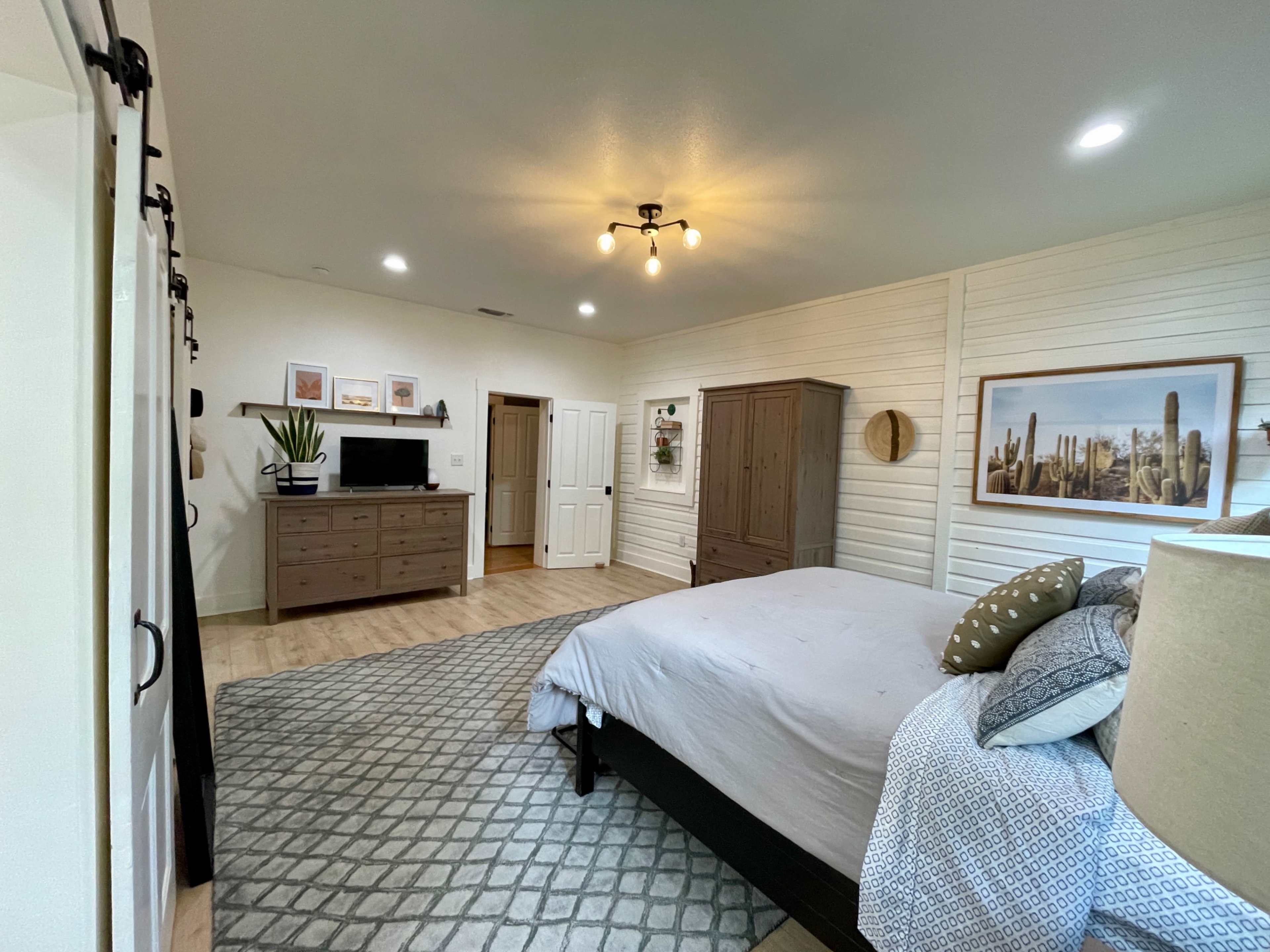 A well-lit bedroom features a bed with decorative pillows, a matching dresser and TV setup, and a textured rug on wooden flooring.
