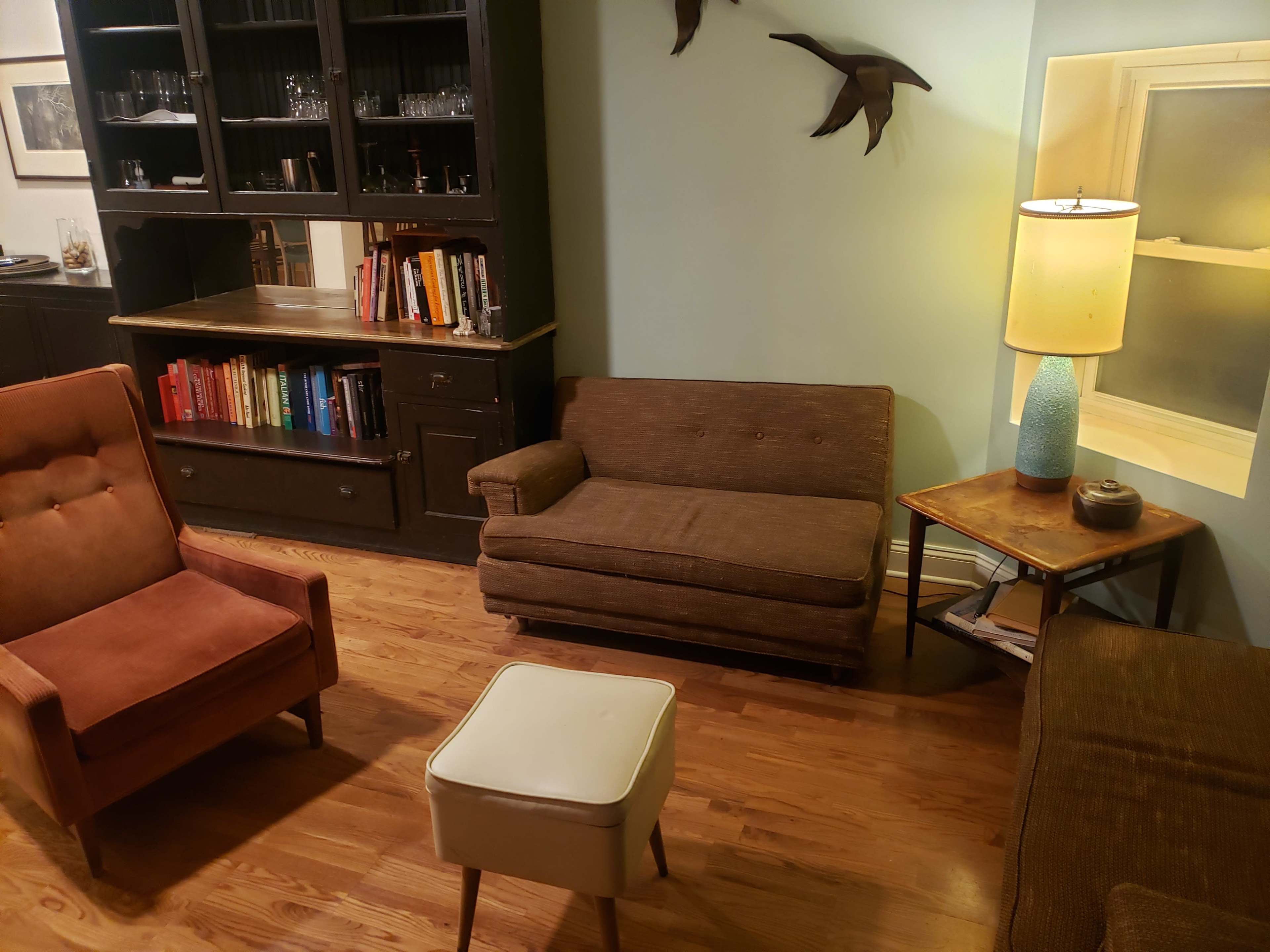 A cozy living room with vintage furniture, including two armchairs, a sofa, a side table with a lamp, and a bookshelf against the wall.
