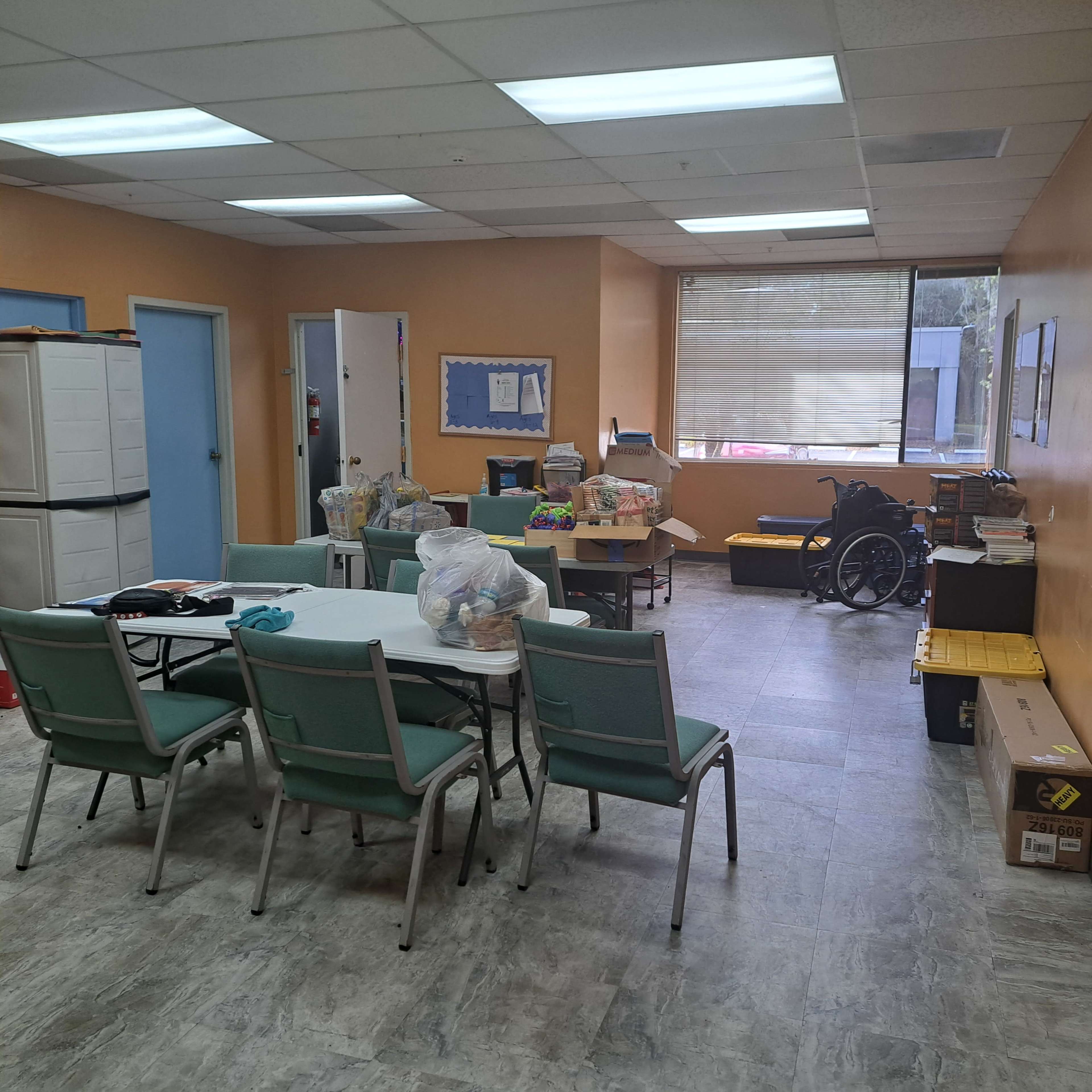 The image shows a brightly lit room with tables and chairs, organized storage units, and various items scattered around, including a wheelchair and boxes.